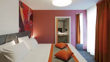 Habitación de hotel con cama, almohadas, mesita de noche, espejo, baño con lavamanos y ventana con cortinas.
