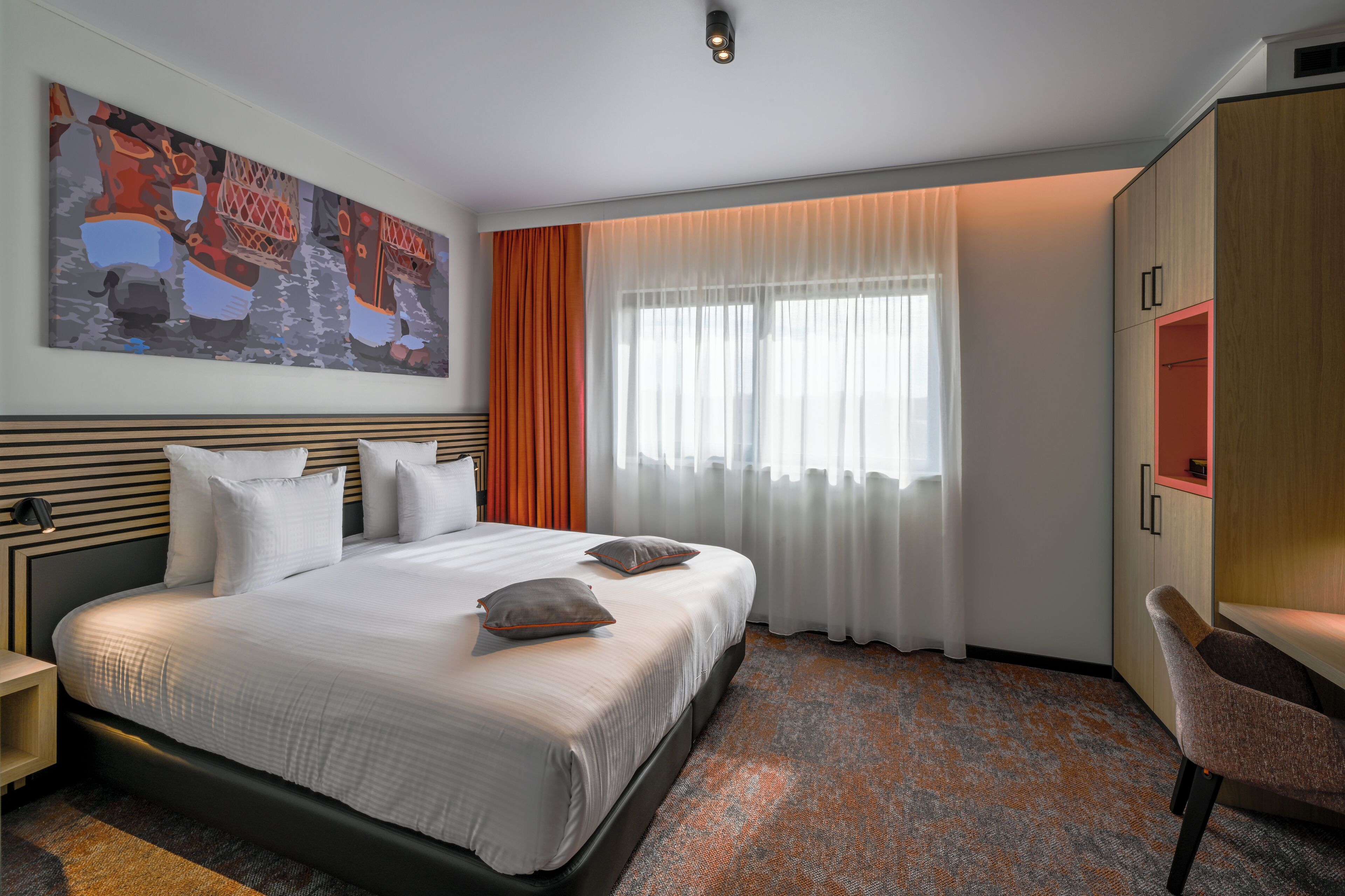 A hotel room with a large bed, a desk, a chair, a wardrobe, and a painting on the wall.