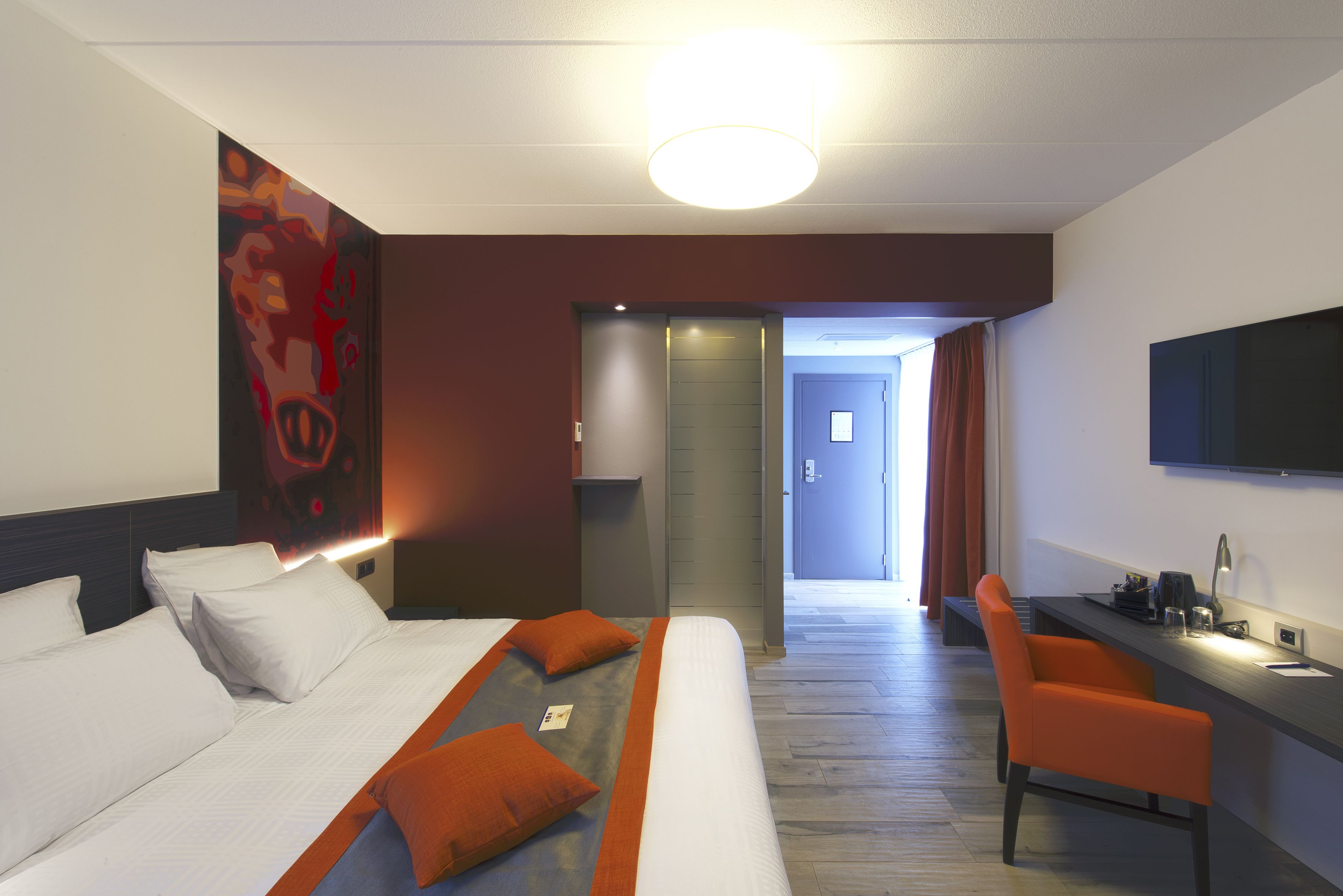 Hotelkamer met een bed, een bureau en een stoel. Er hangt een schilderij aan de muur, een televisie is aan de muur bevestigd en er is een deur naar een andere kamer.