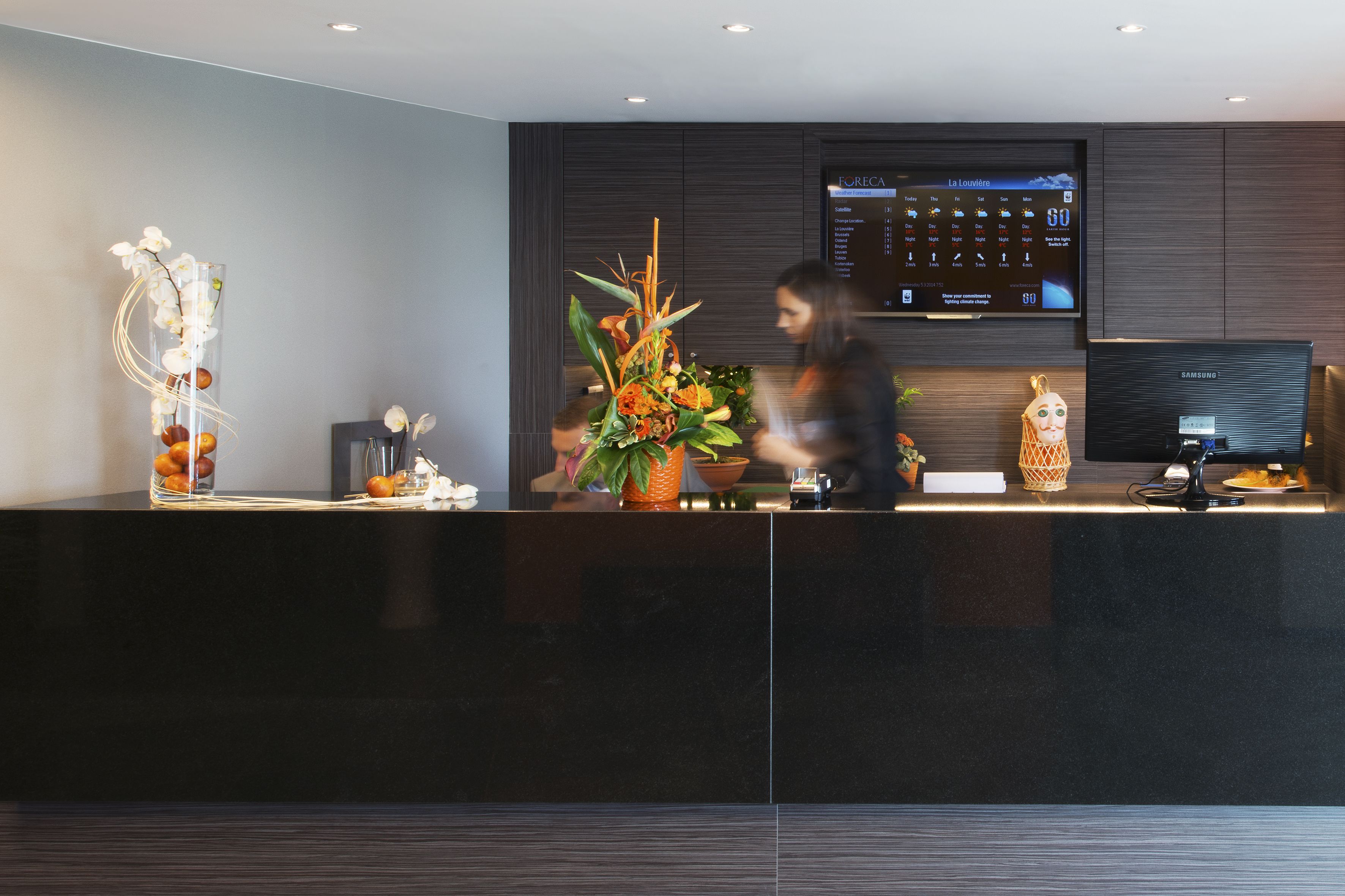 Una moderna área de recepción con una gran pantalla digital, un mostrador de recepción y plantas decorativas.