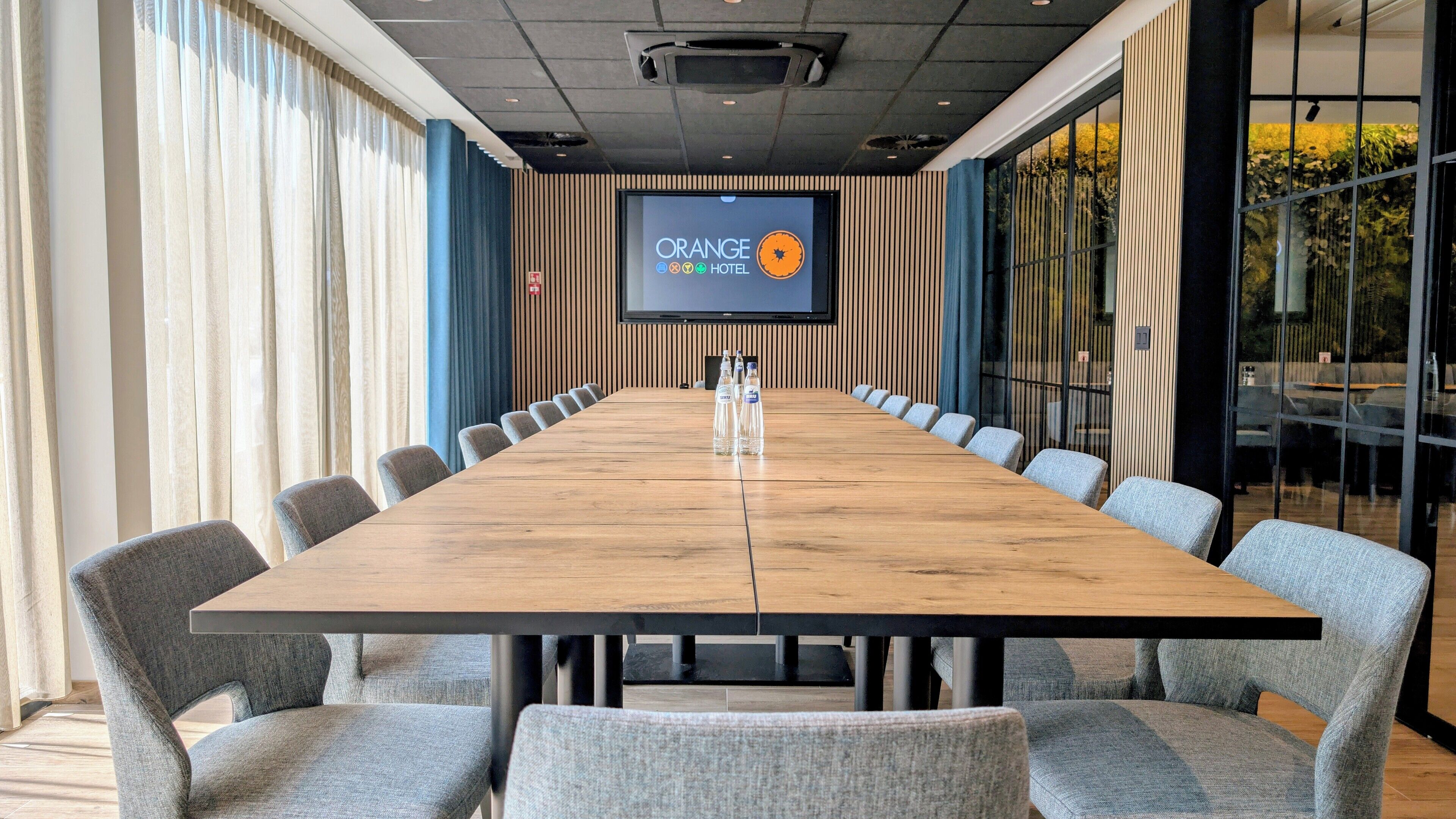 A conference room with a long wooden table, grey upholstered chairs, and a large screen displaying the Orange Hotel logo.