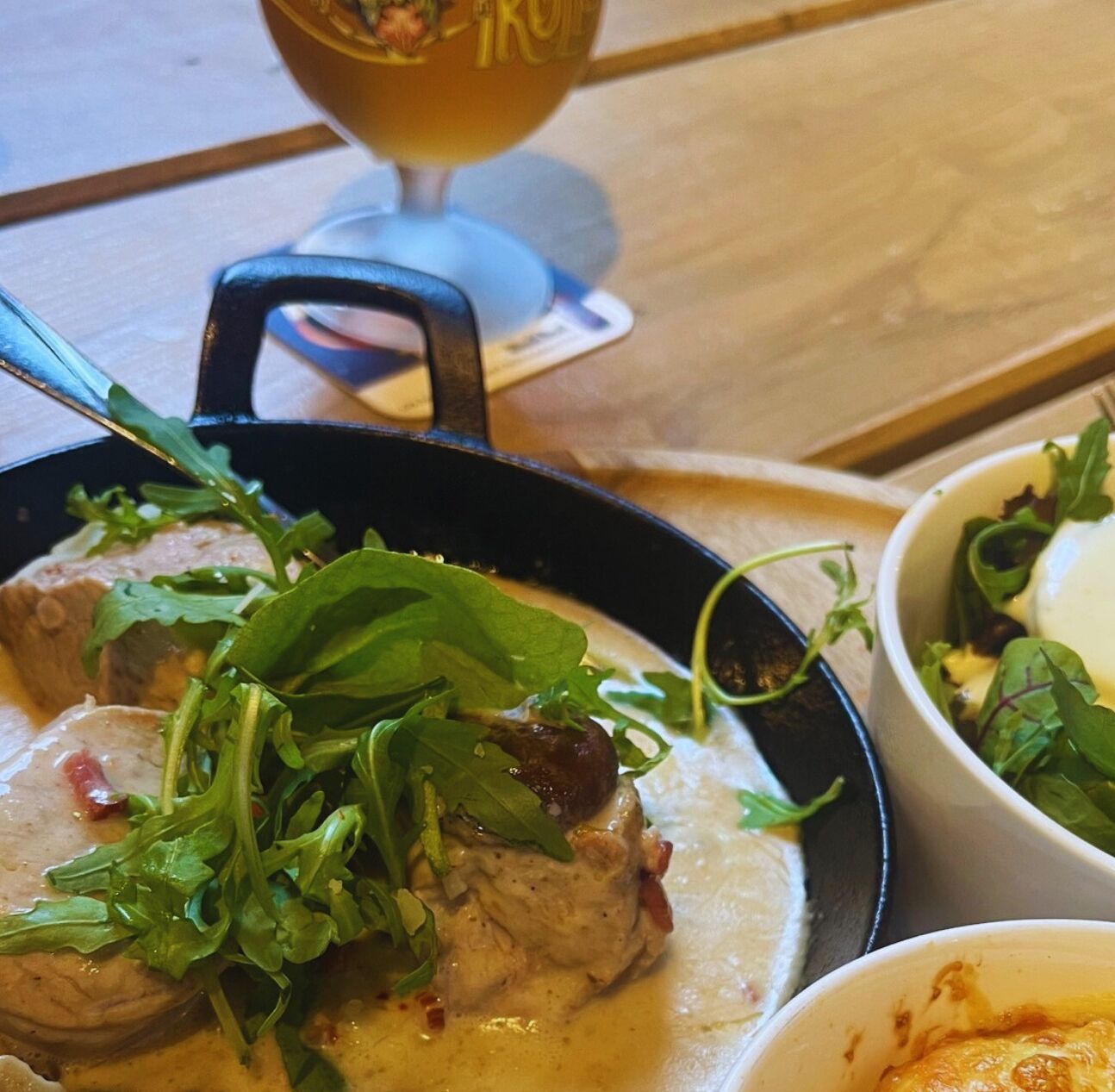 A meal with meat and greens in a pan, a glass of beer, and a bowl of salad on a wooden table.
