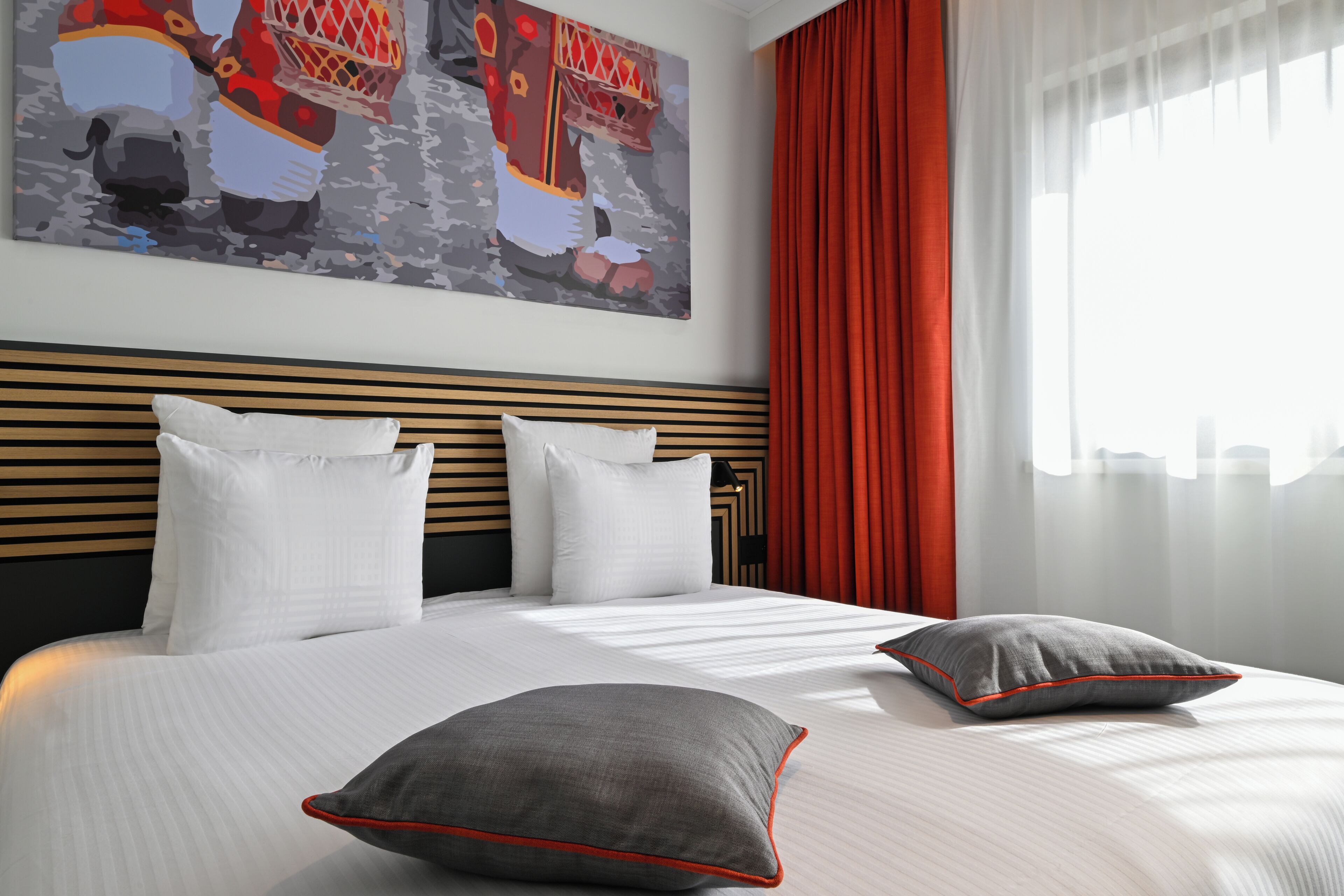 Een hotelkamer met een groot bed, een hoofdbord met horizontale latten, een schilderij boven het bed en rode gordijnen.