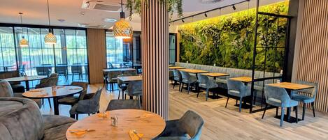 A modern restaurant with round tables, blue chairs, and a green wall with plants.