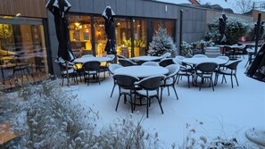 Un espace extérieur aménagé avec des tables et des chaises, recouvert de neige, un petit sapin de Noël et un bâtiment doté de grandes fenêtres.