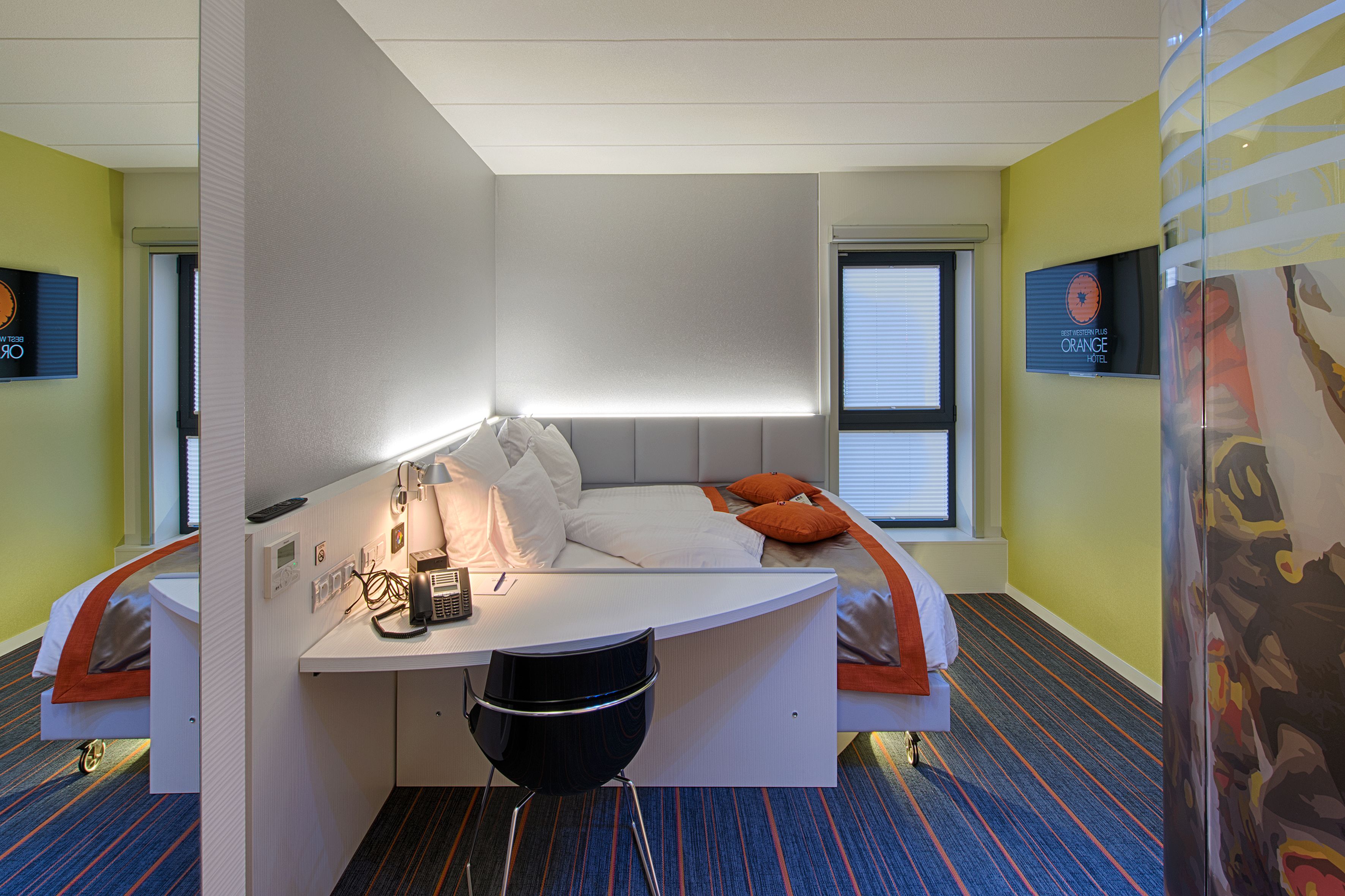A modern hotel room with a curved desk, a bed with orange pillows, and a view of the city through the window.