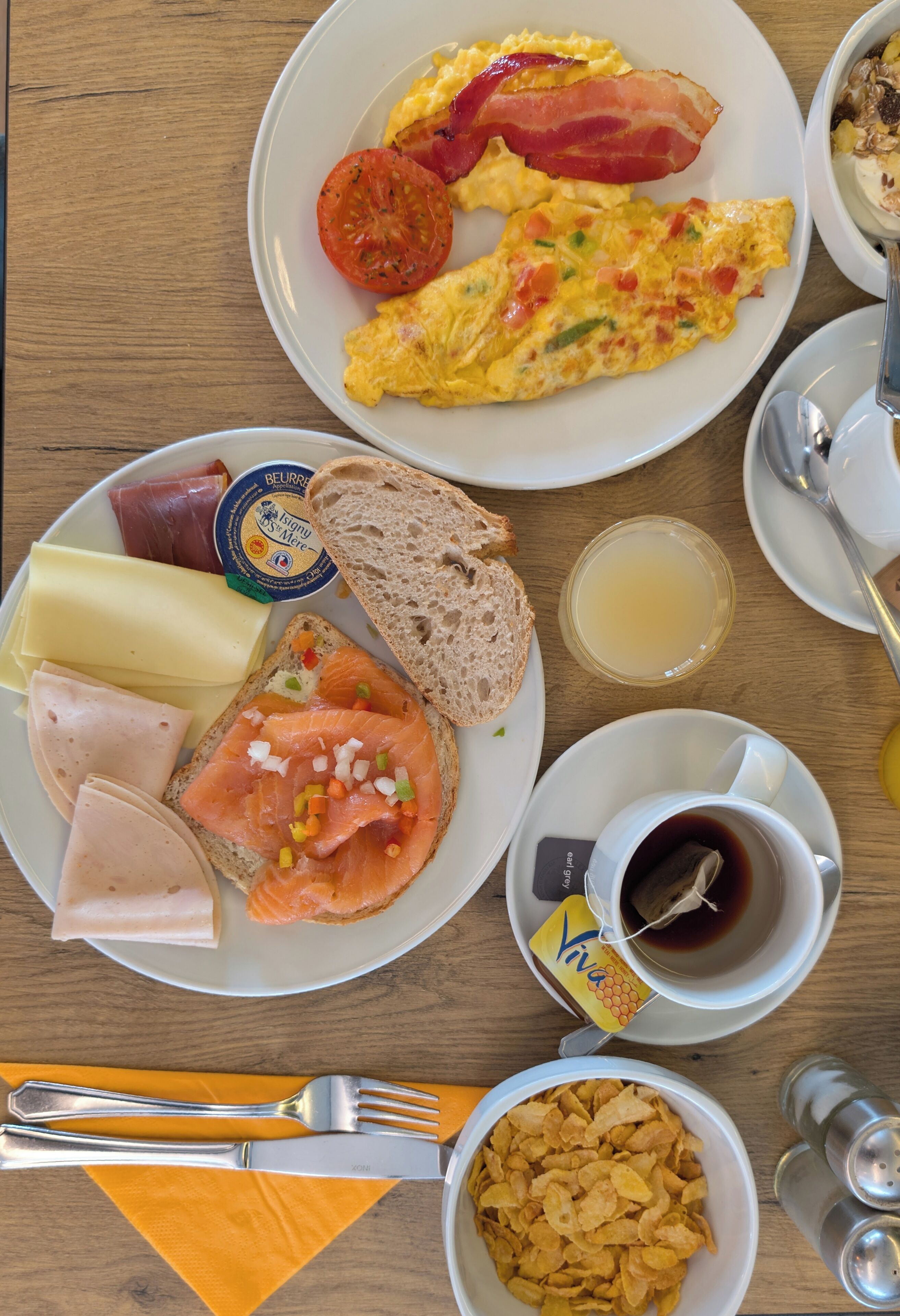 Una mesa de desayuno con platos de huevos, tocino, jamón, queso, salmón ahumado, tostadas y una taza de té.