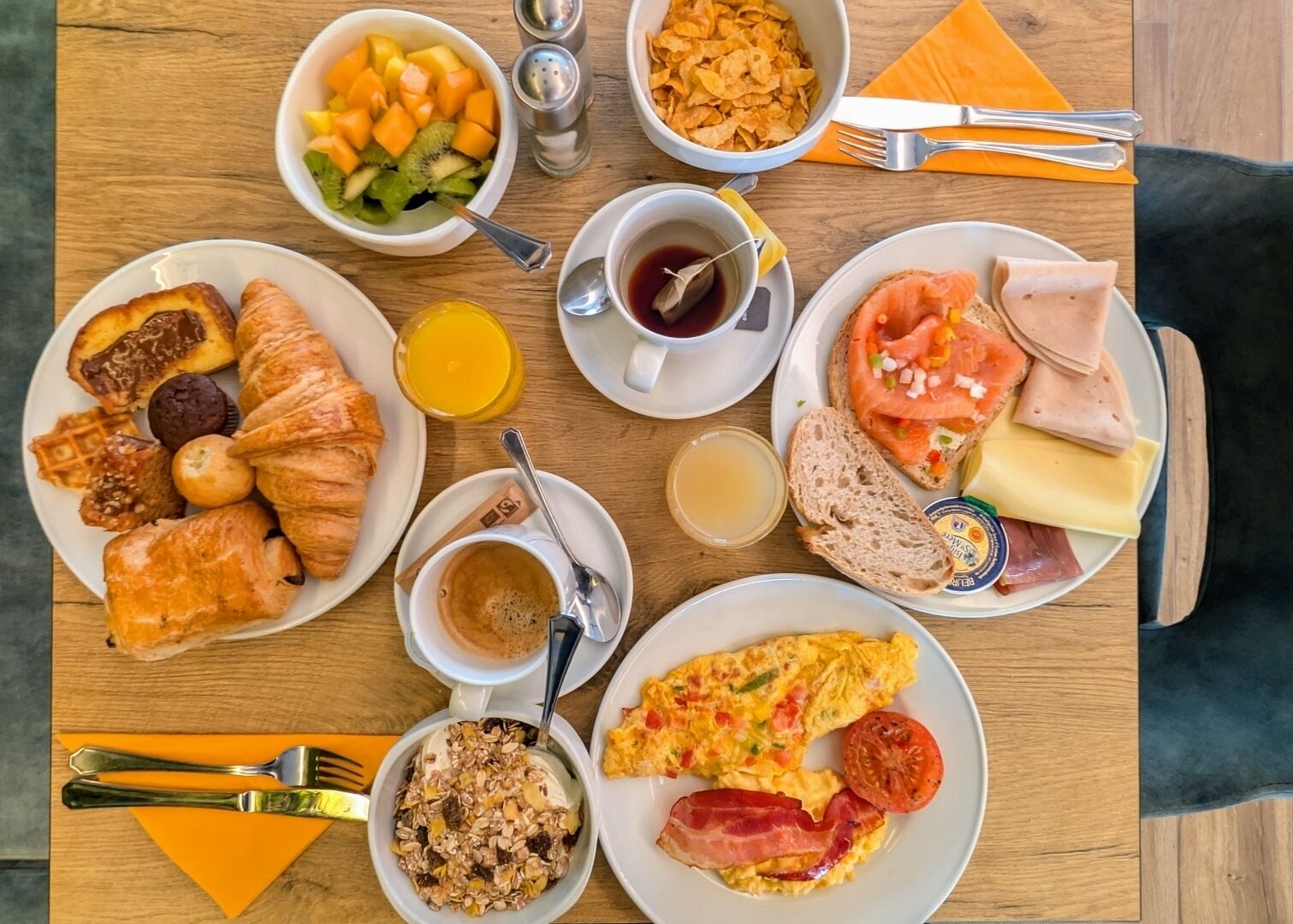 A breakfast table with croissants, eggs, ham, smoked salmon, and various fruits and cereals.
