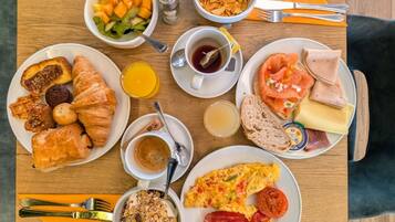 A breakfast table with croissants, eggs, ham, smoked salmon, and various fruits and cereals.