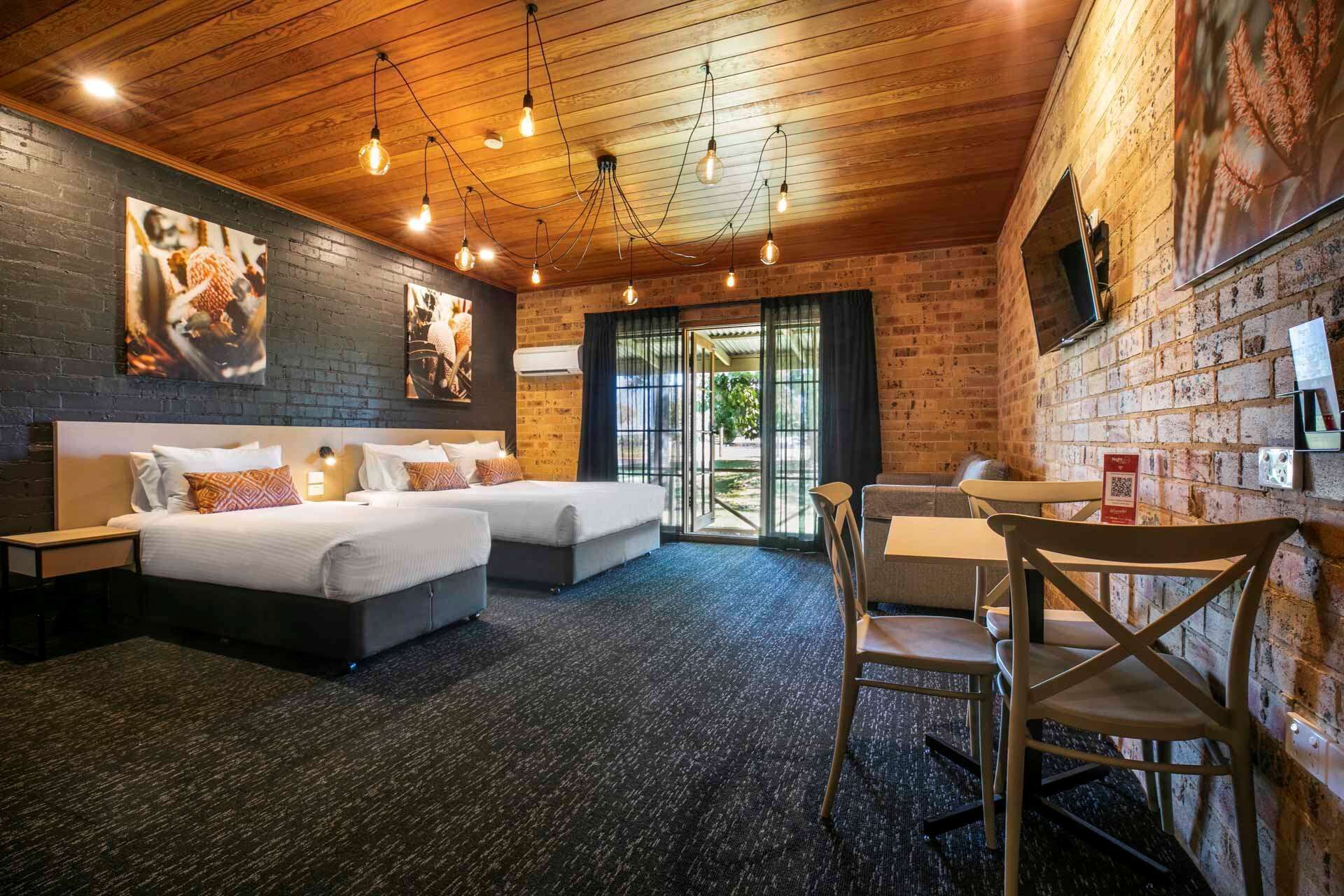 A hotel room with two beds, a desk, and a chair. There are light fixtures on the ceiling and a brick wall.