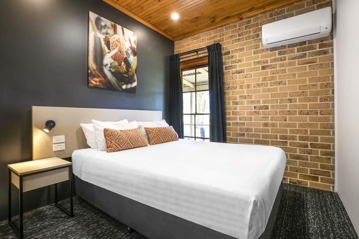 A hotel room with a large bed, a nightstand, a wall-mounted air conditioner, and a brick wall.