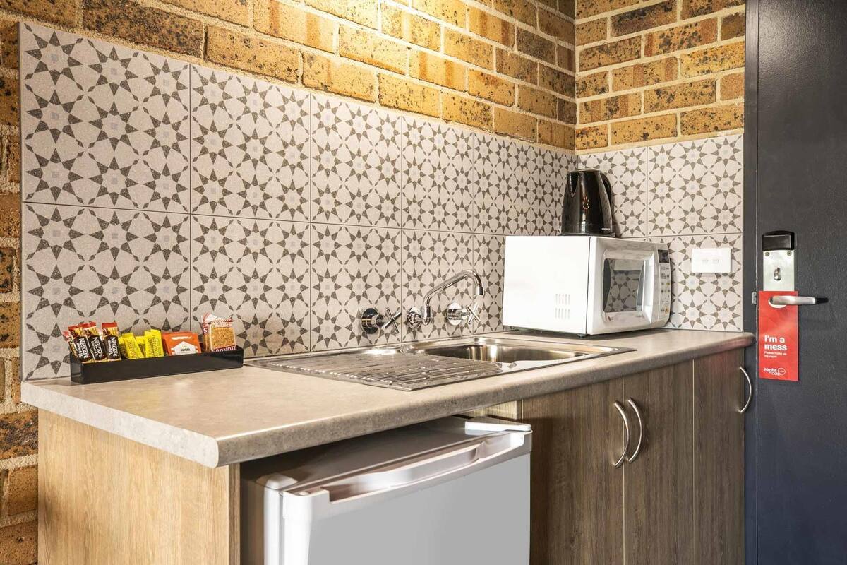A kitchen with a microwave, sink, and coffee maker. There is a shelf with condiments and a door with a sign.