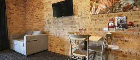 A room with a brick wall, a flat-screen TV mounted above a table and chairs, a sofa, and a poster on the wall.