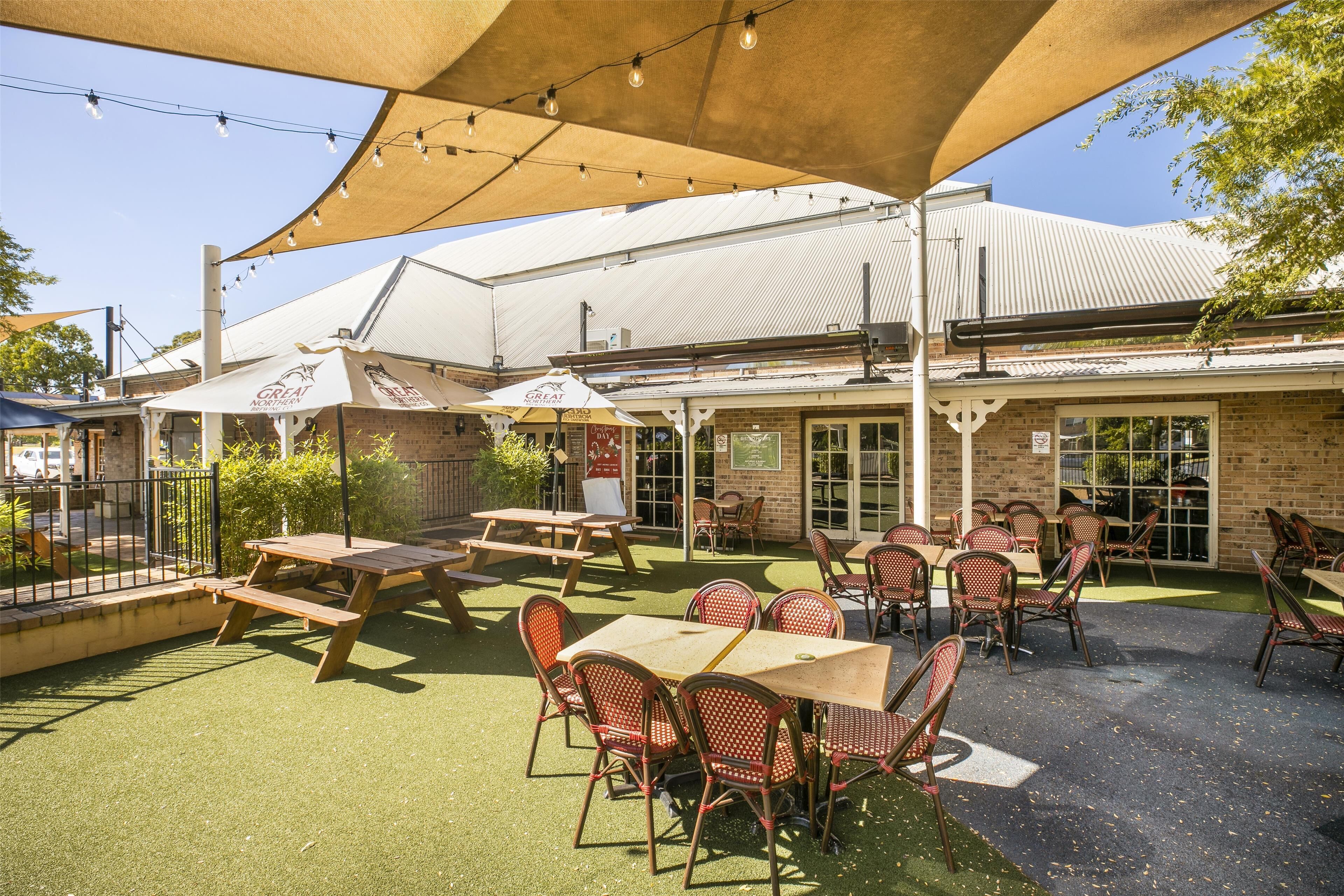 An outdoor patio area with tables and chairs, a covered seating area, and a brick building in the background.