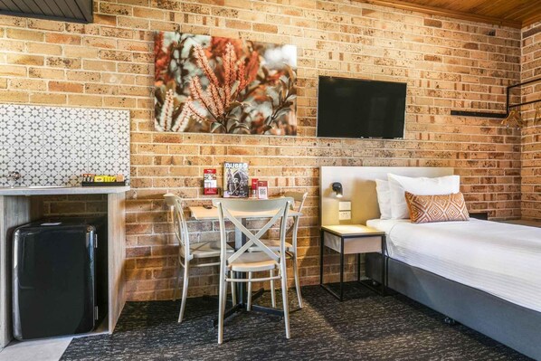 A compact hotel room with a brick wall, a bed, a desk with a chair, a small table, a TV, and a trash bin.