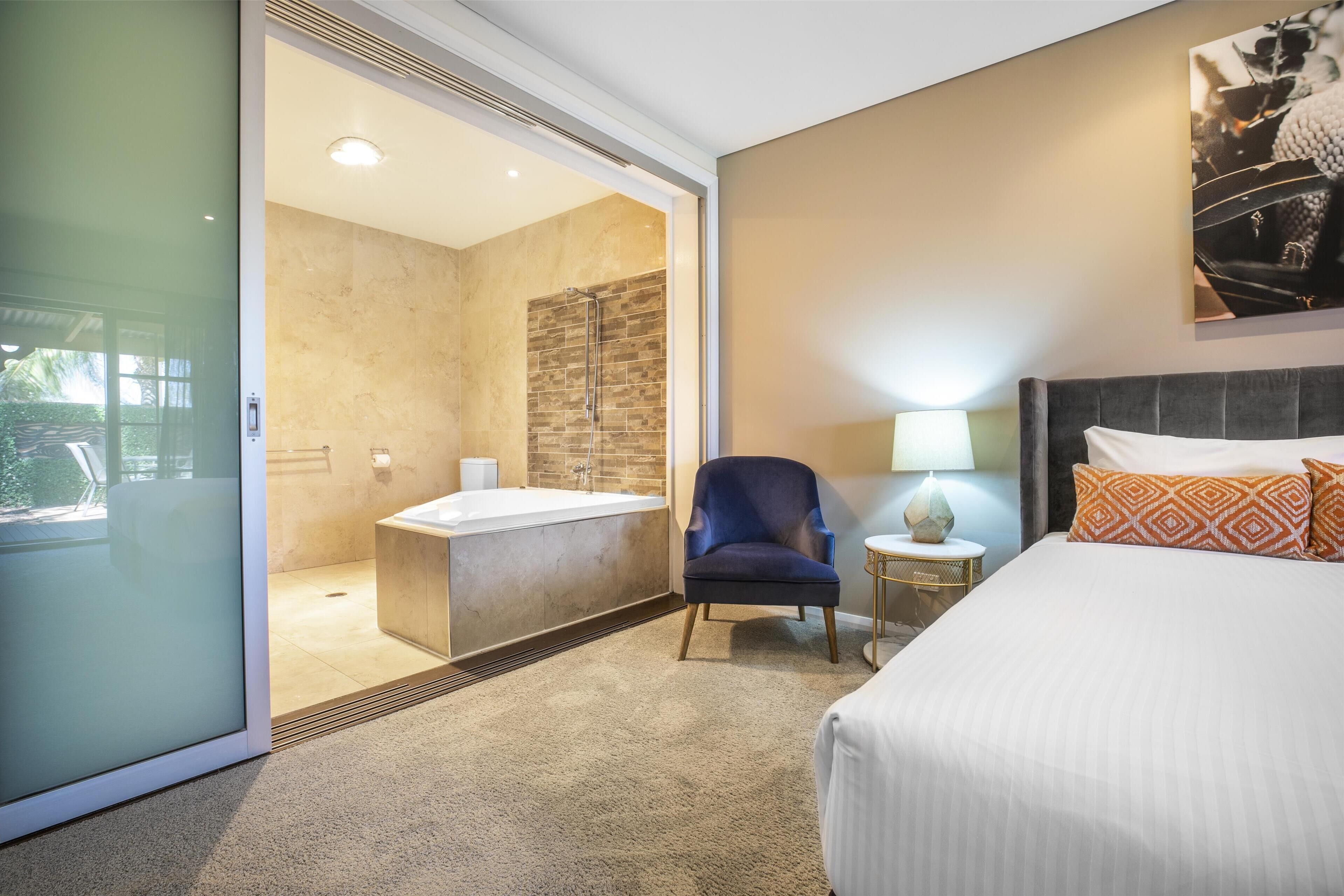 A hotel room with a large bed, a sitting area with a chair, a small round table, a wall-mounted lamp, and a bathroom with a bathtub and shower.