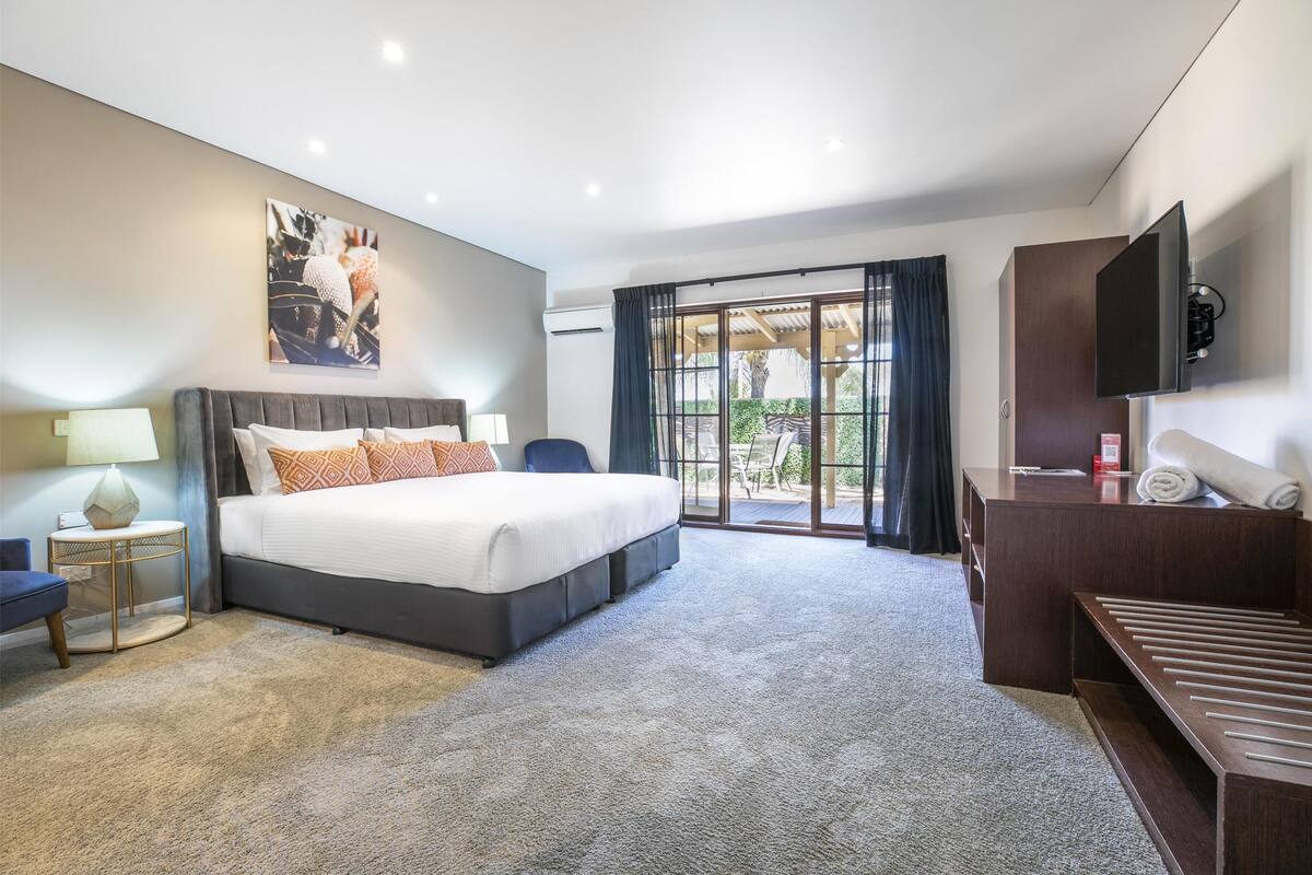 A modern hotel room with a large bed, a desk, a TV, and a balcony with a view.