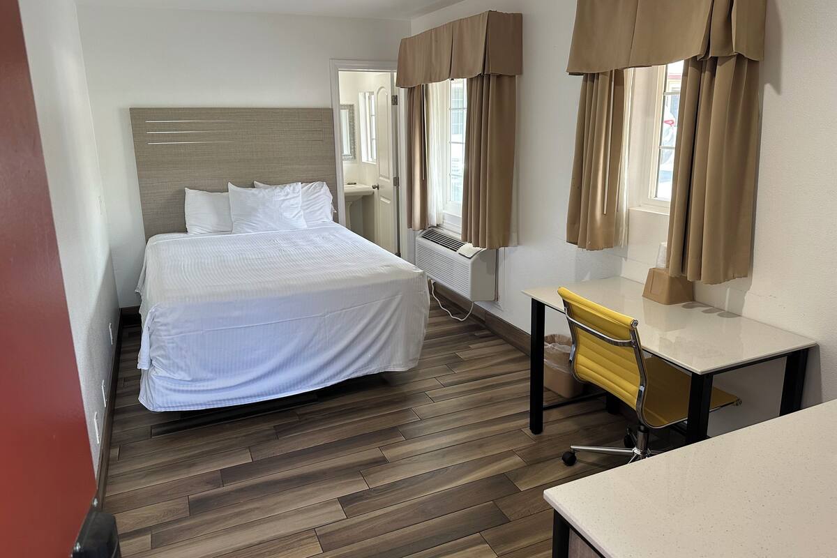 A hotel room with a bed, a desk, and a chair. There is a window with curtains, a radiator, and a door leading to another room.