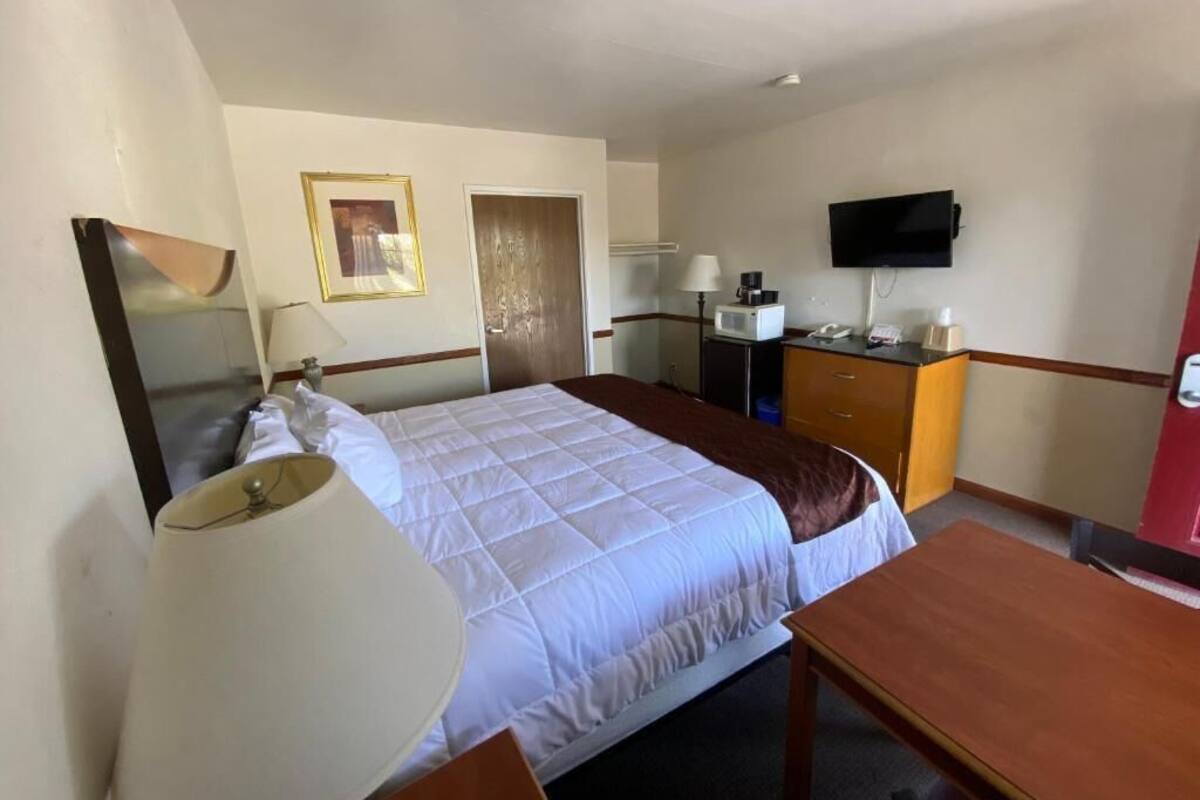 A hotel room with a bed, bedside table, lamp, TV, and a desk with a chair.