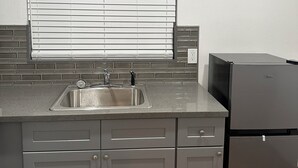 A kitchen with a stainless steel sink, a black refrigerator, grey cabinets, a window with blinds, and a tiled backsplash.