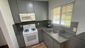 A compact kitchen with grey cabinets, a white stove, a microwave, a sink, and a window with blinds.