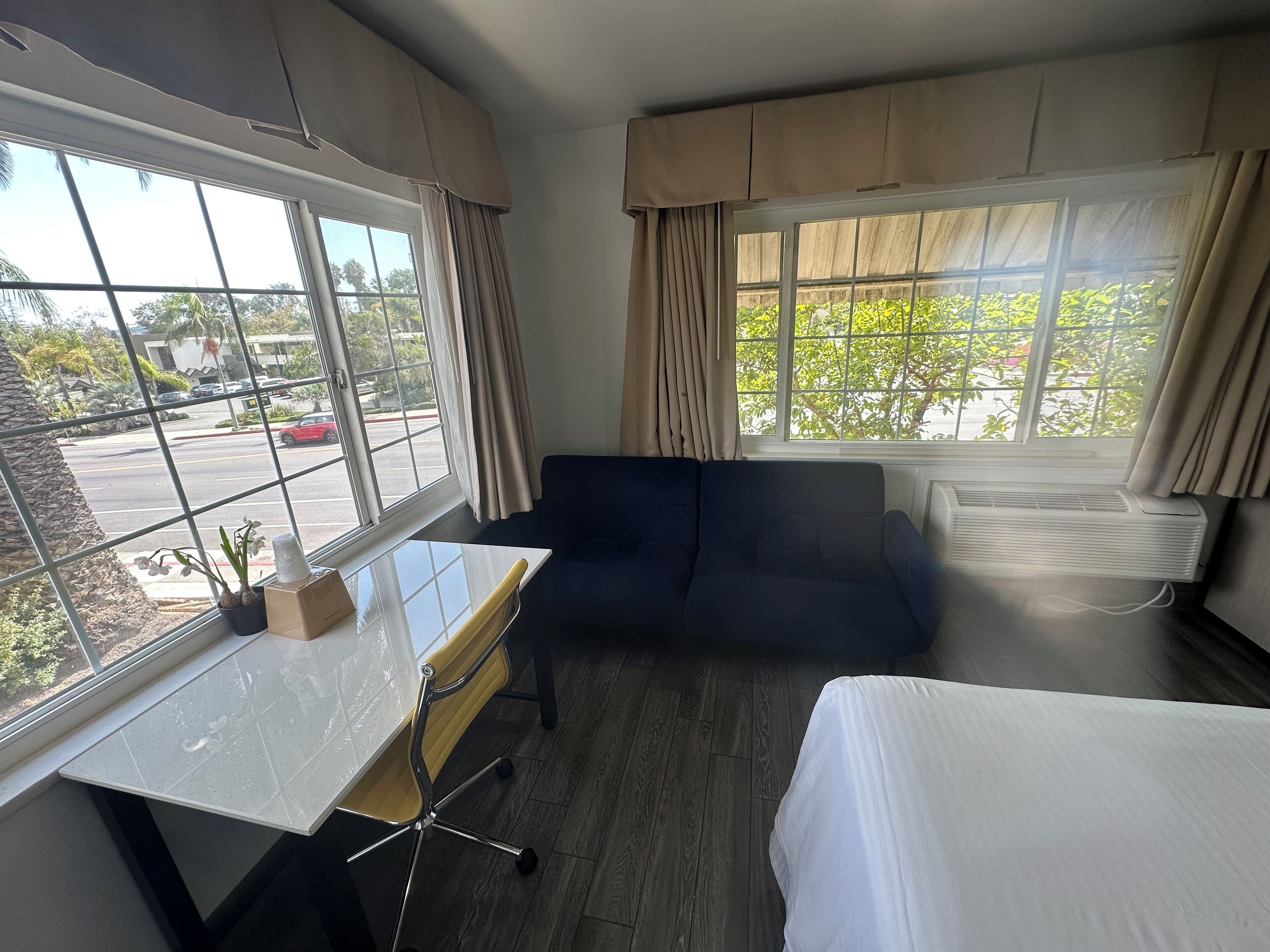 Habitació d'hotel amb llit, escriptori, cadira, sofà i dues finestres amb cortines.