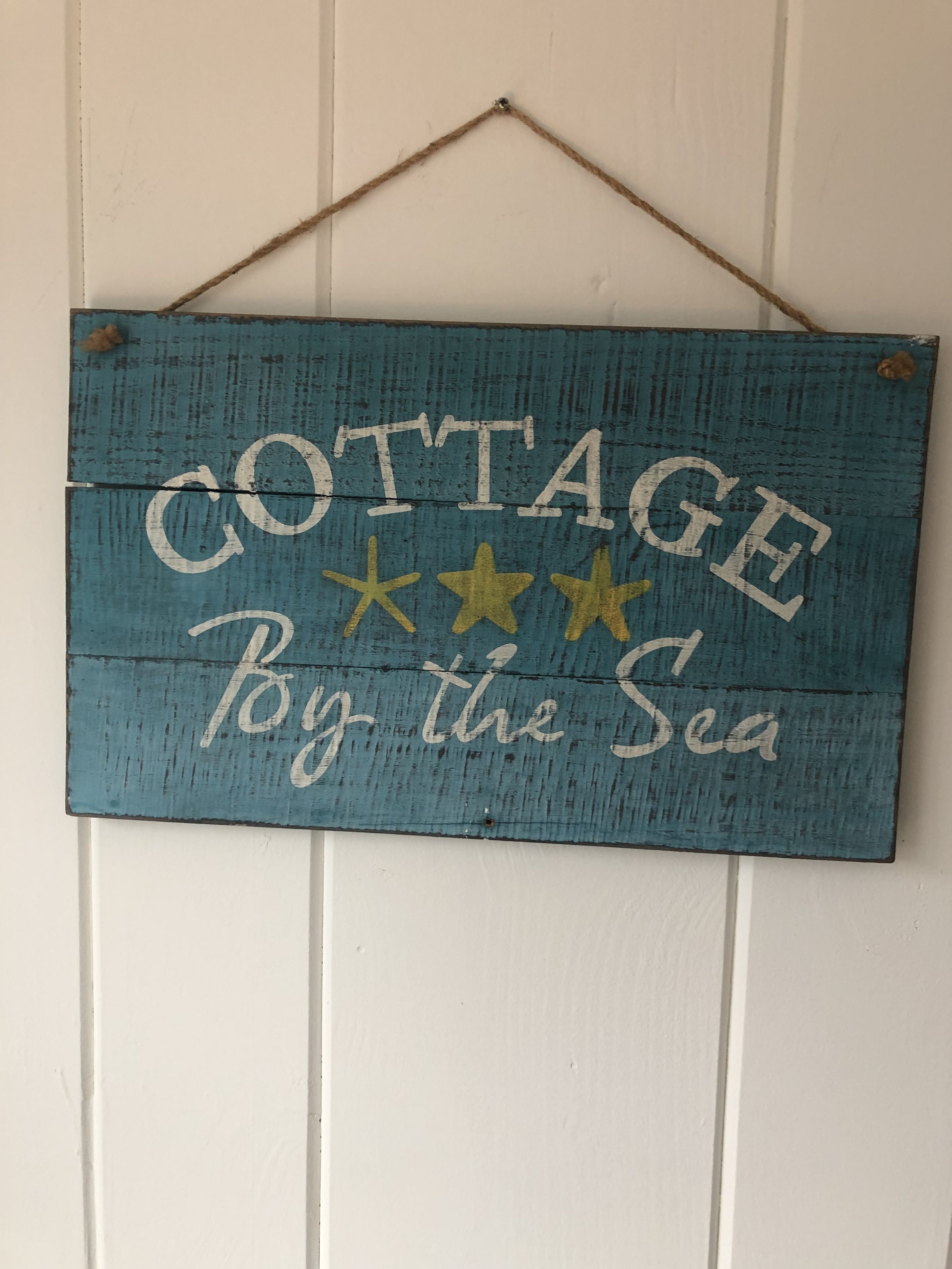 A rustic sign with the text "COTTAGE By the Sea" hanging on a white wall.