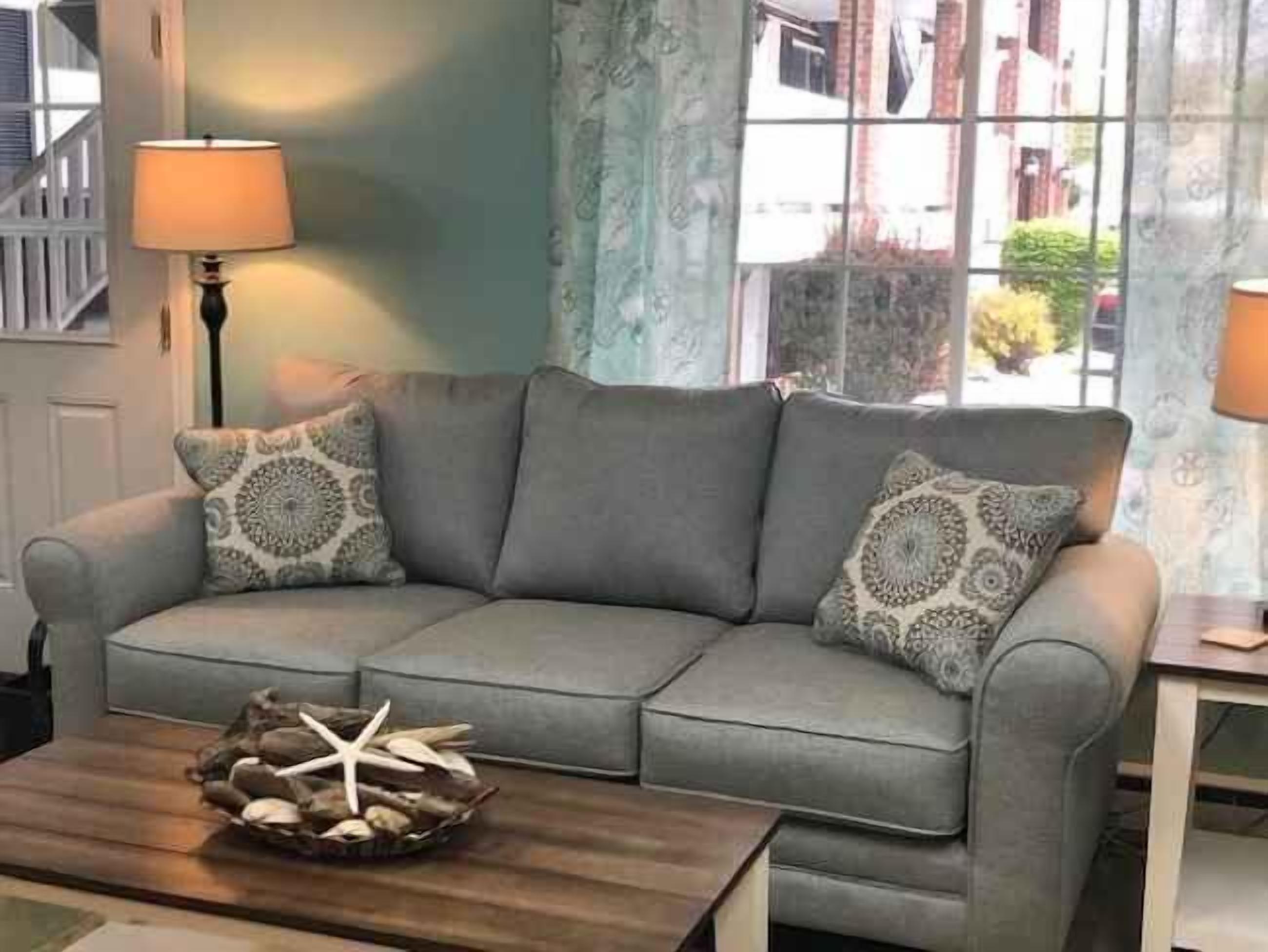 A grey sofa with patterned cushions, a wooden coffee table with starfish and shells, and a lamp on a side table.