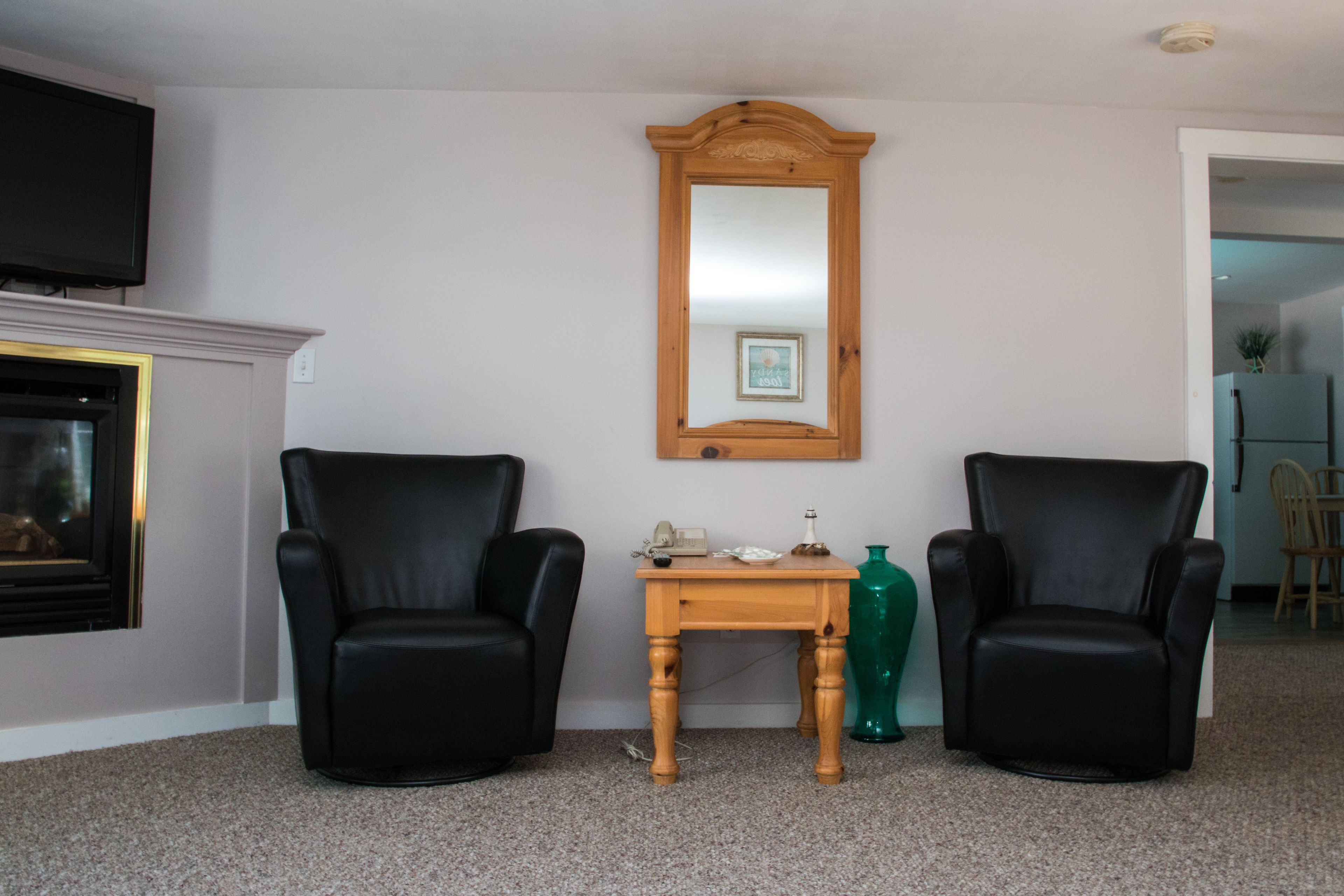 A living room with two black leather chairs, a wooden table with a telephone and decorative items, a mirror on the wall, and a television mounted above a fireplace.