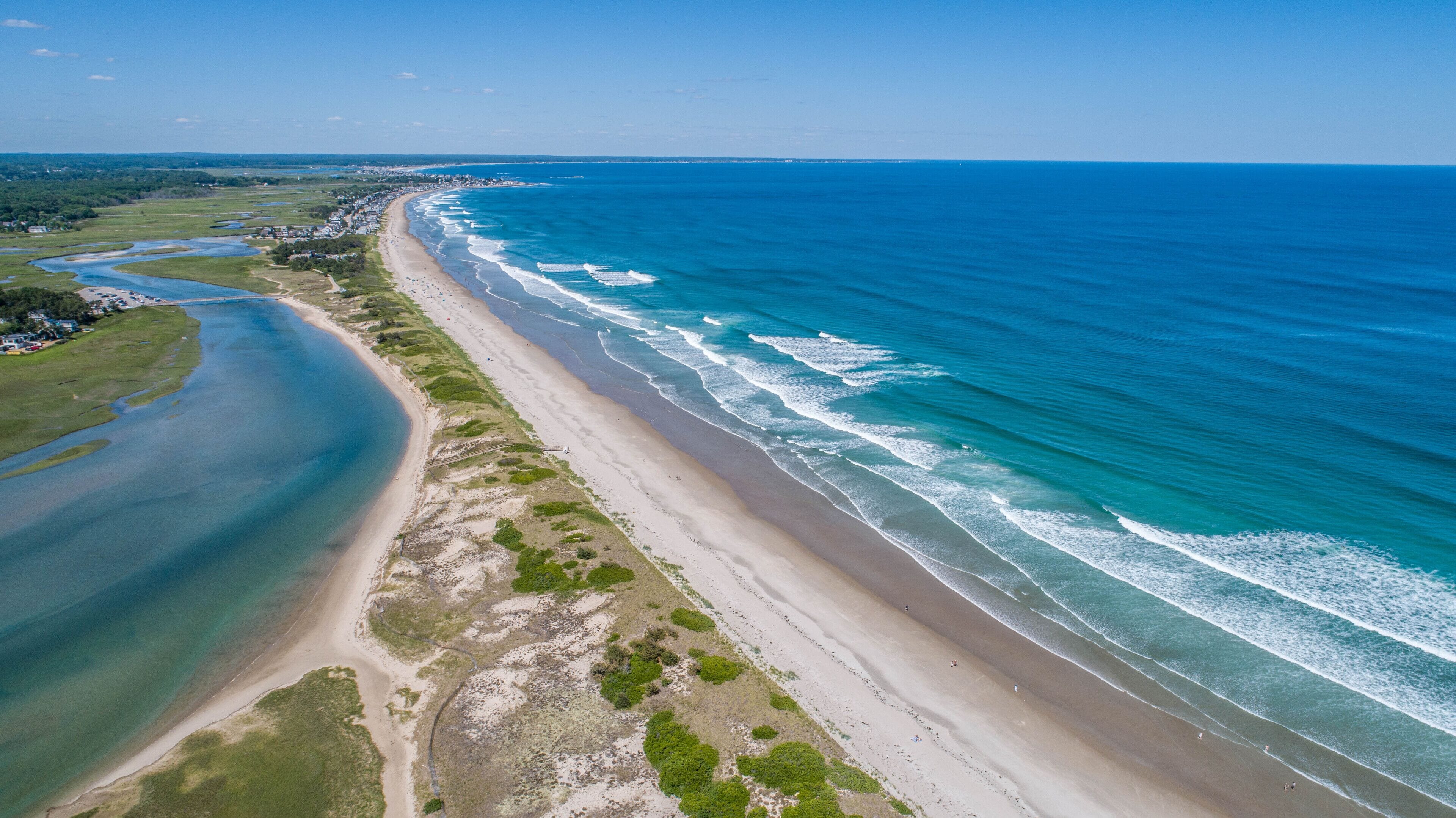 An aerial view of a coastal area with a long sandy beach, a river or bay to the left, and a clear blue ocean to the right.