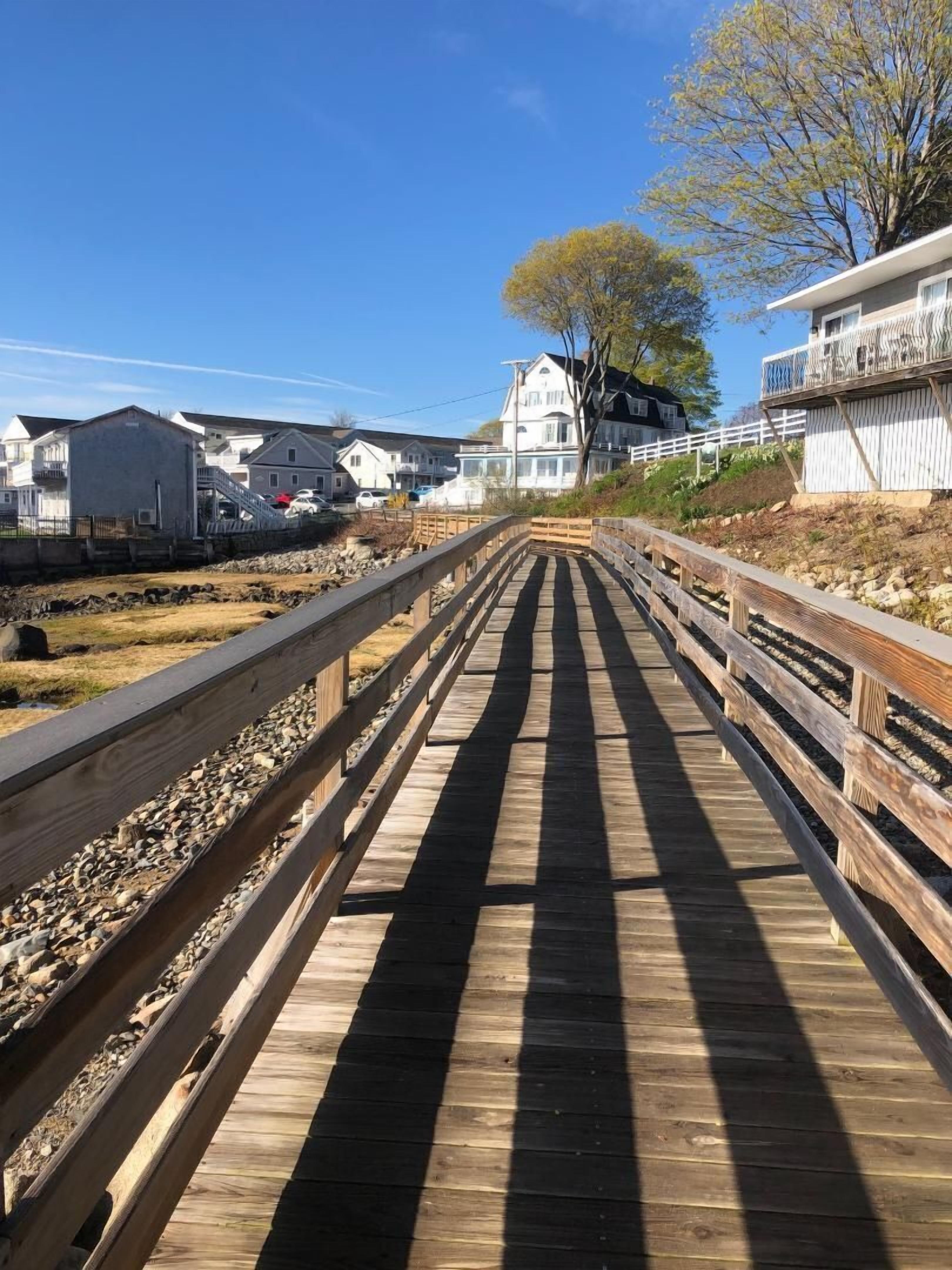 En træpromenade der fører til en strand med huse på den ene side og et stort træ på den anden.