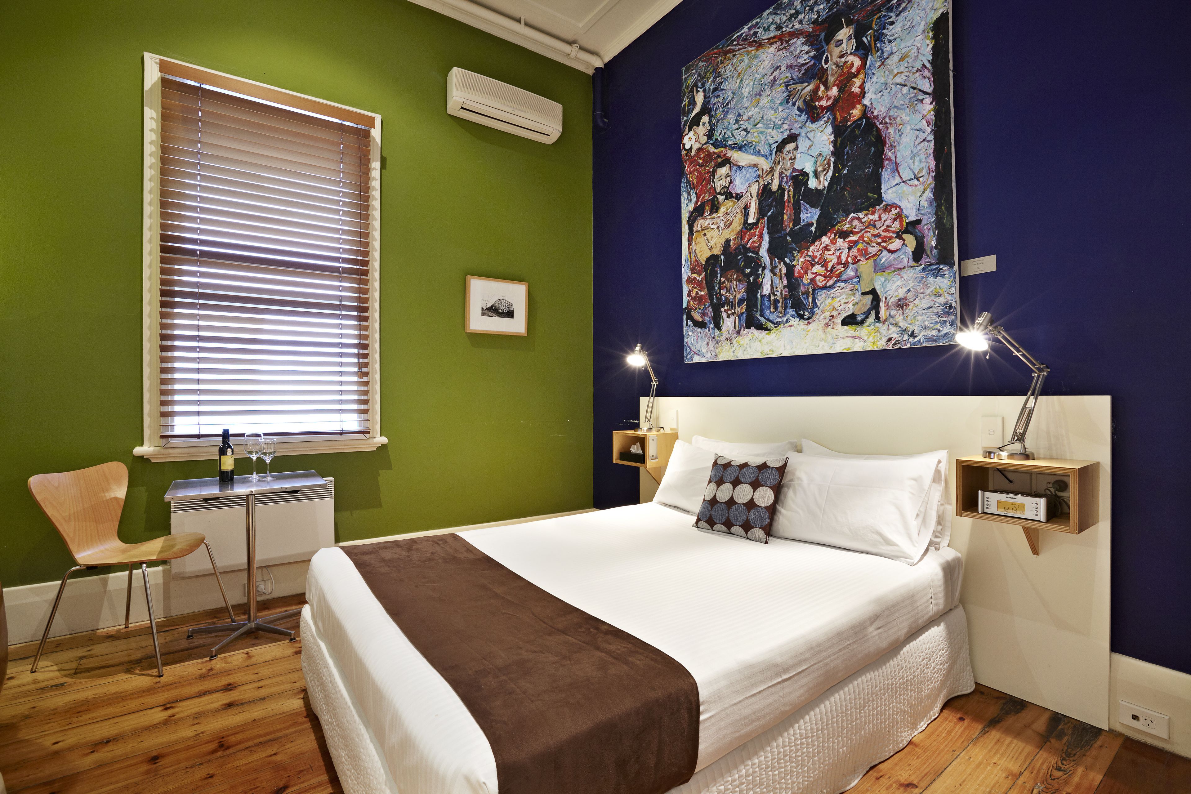 Una habitación de hotel con cama, escritorio, silla, un cuadro en la pared y ventana con persianas.