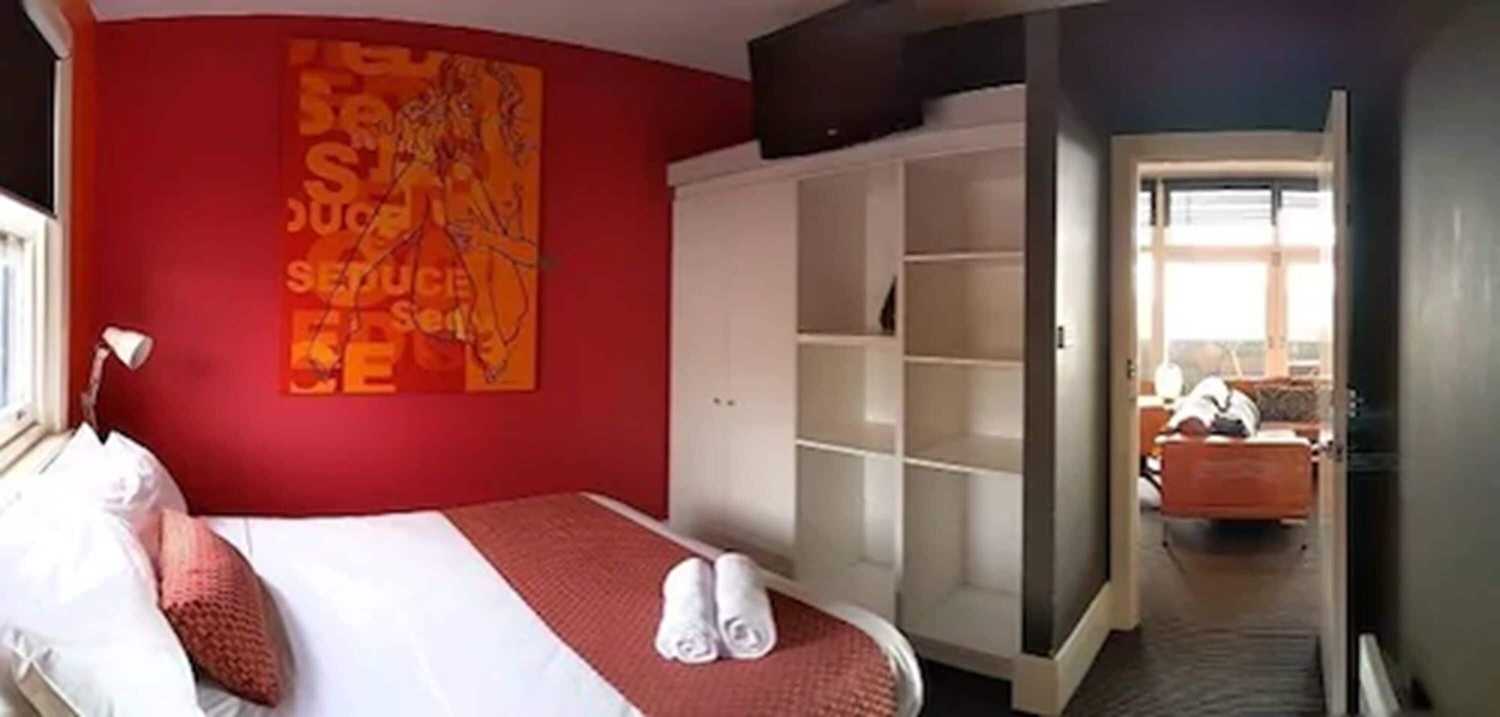 A hotel room with a red accent wall, a bed with white linens, a large wardrobe, and a view through an open door.