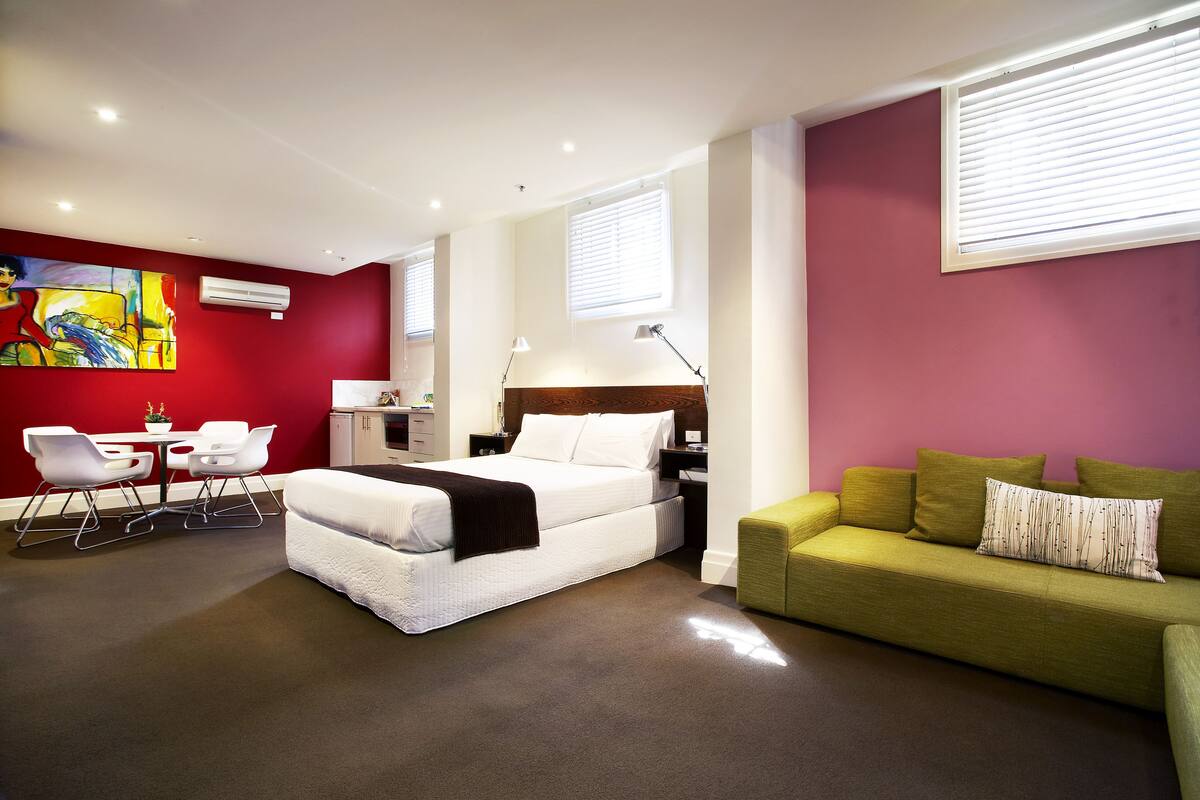 A modern hotel room with a bed, a green sofa, a desk with a chair, and a painting on the wall.