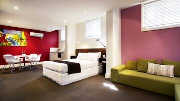 A modern hotel room with a bed, a green sofa, a desk with a chair, and a painting on the wall.