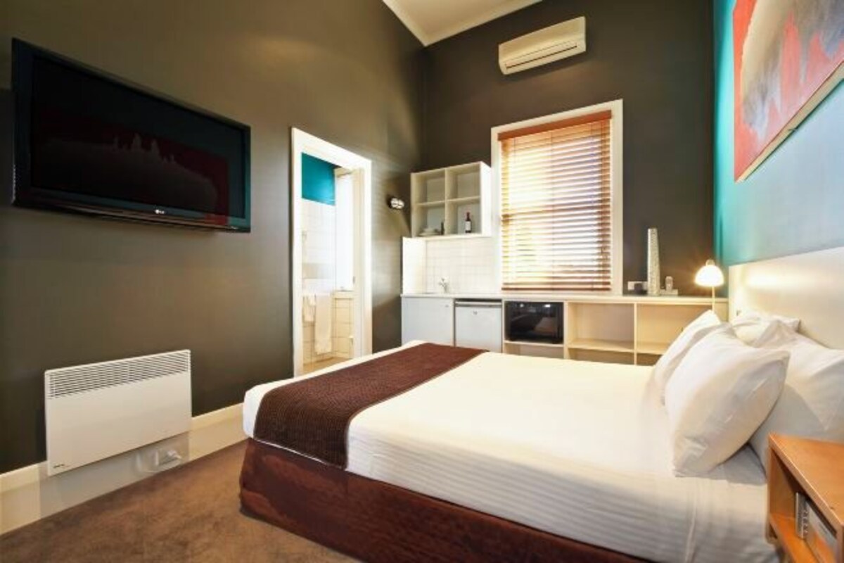 A hotel room with a bed, a television, a desk, a window with blinds, and a kitchenette.
