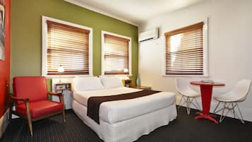 Quarto de hotel com cama, uma poltrona vermelha, uma mesinha e duas cadeiras brancas.