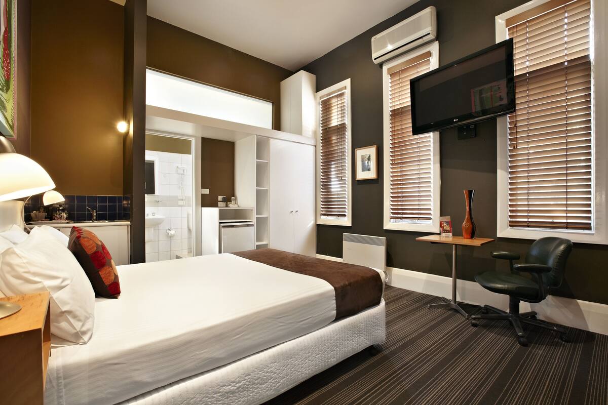 A hotel room with a bed, a desk, a chair, a TV, and a window with blinds.