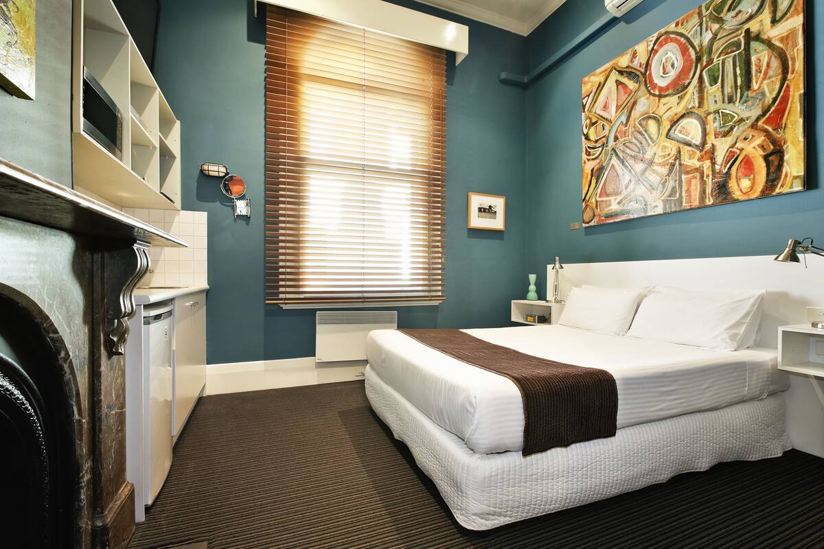 A hotel room with a bed, a dresser, a window with blinds, a painting on the wall, and a radiator.