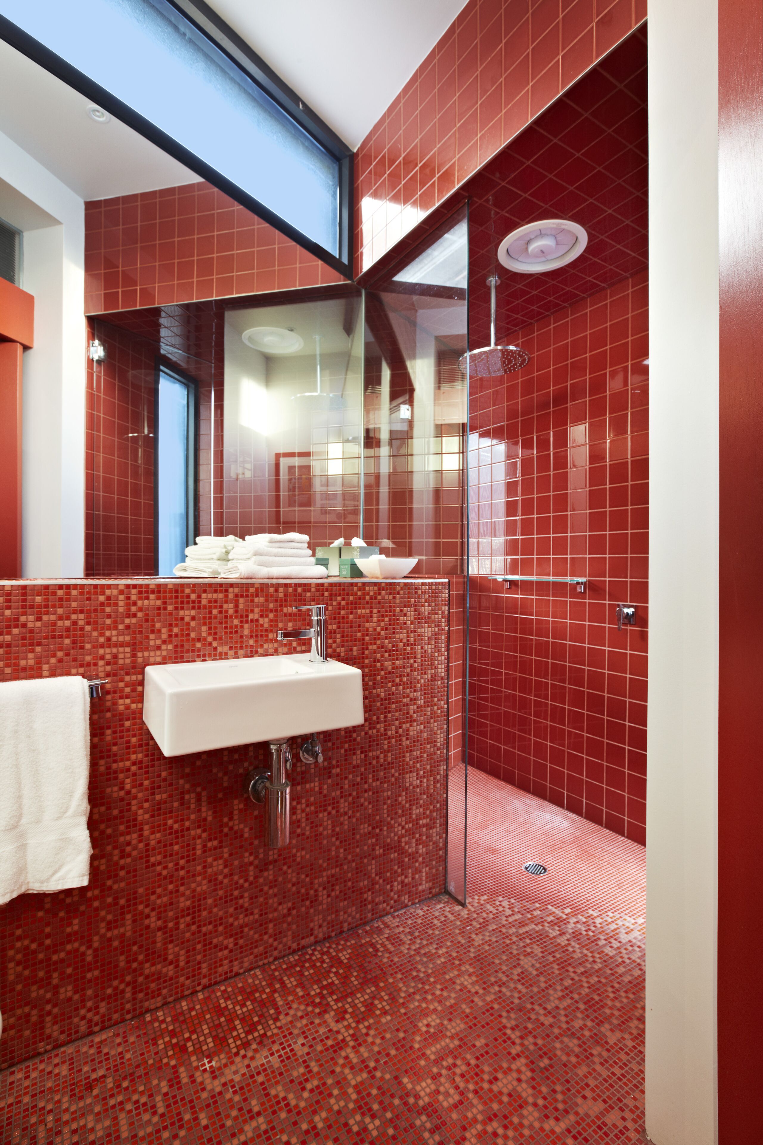 Uma casa de banho moderna com chuveiro em azulejo vermelho, área de chuveiro com divisória de vidro e uma pia branca com espelho em cima.