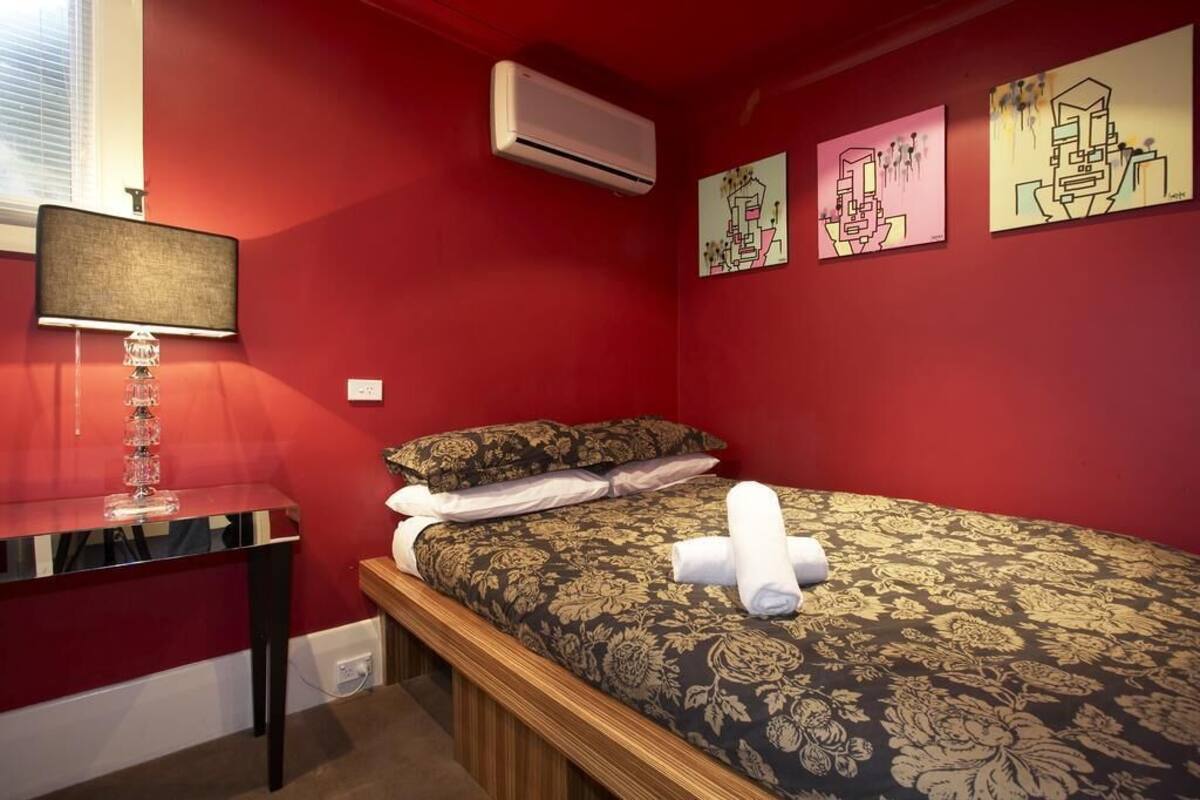 A hotel room with a red wall, a bed with floral bedding, a bedside table with a lamp, and artwork on the wall.