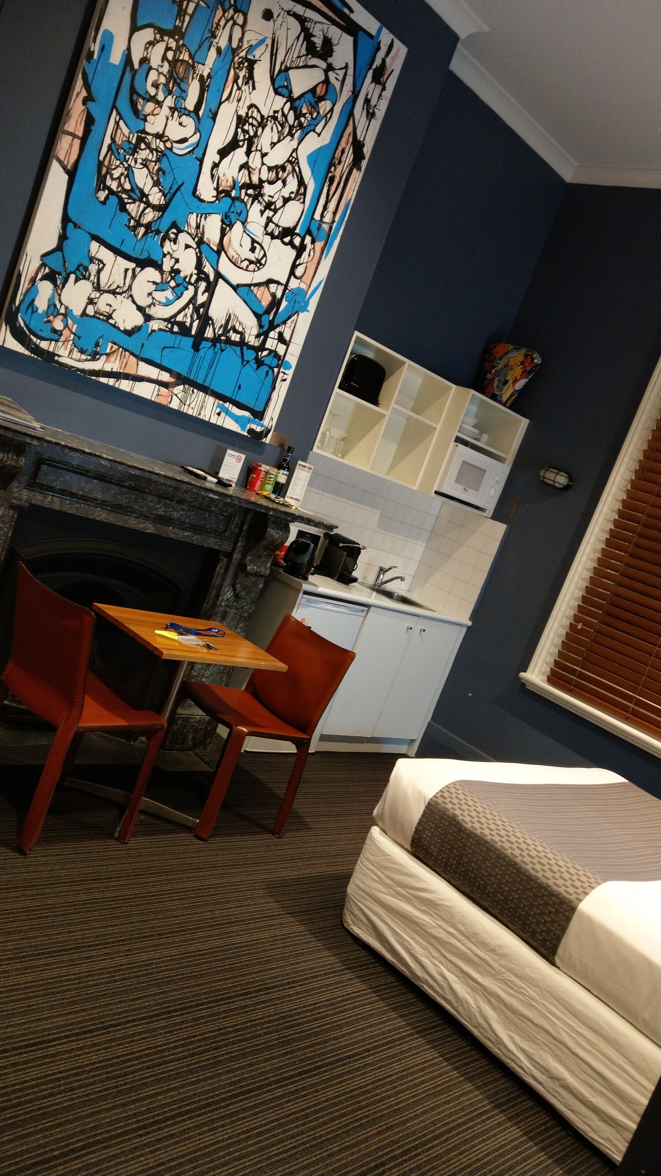 A hotel room with a bed, a dining table with chairs, a kitchen area, and a large abstract painting on the wall.