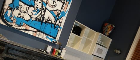 A hotel room with a bed, a dining table with chairs, a kitchen area, and a large abstract painting on the wall.