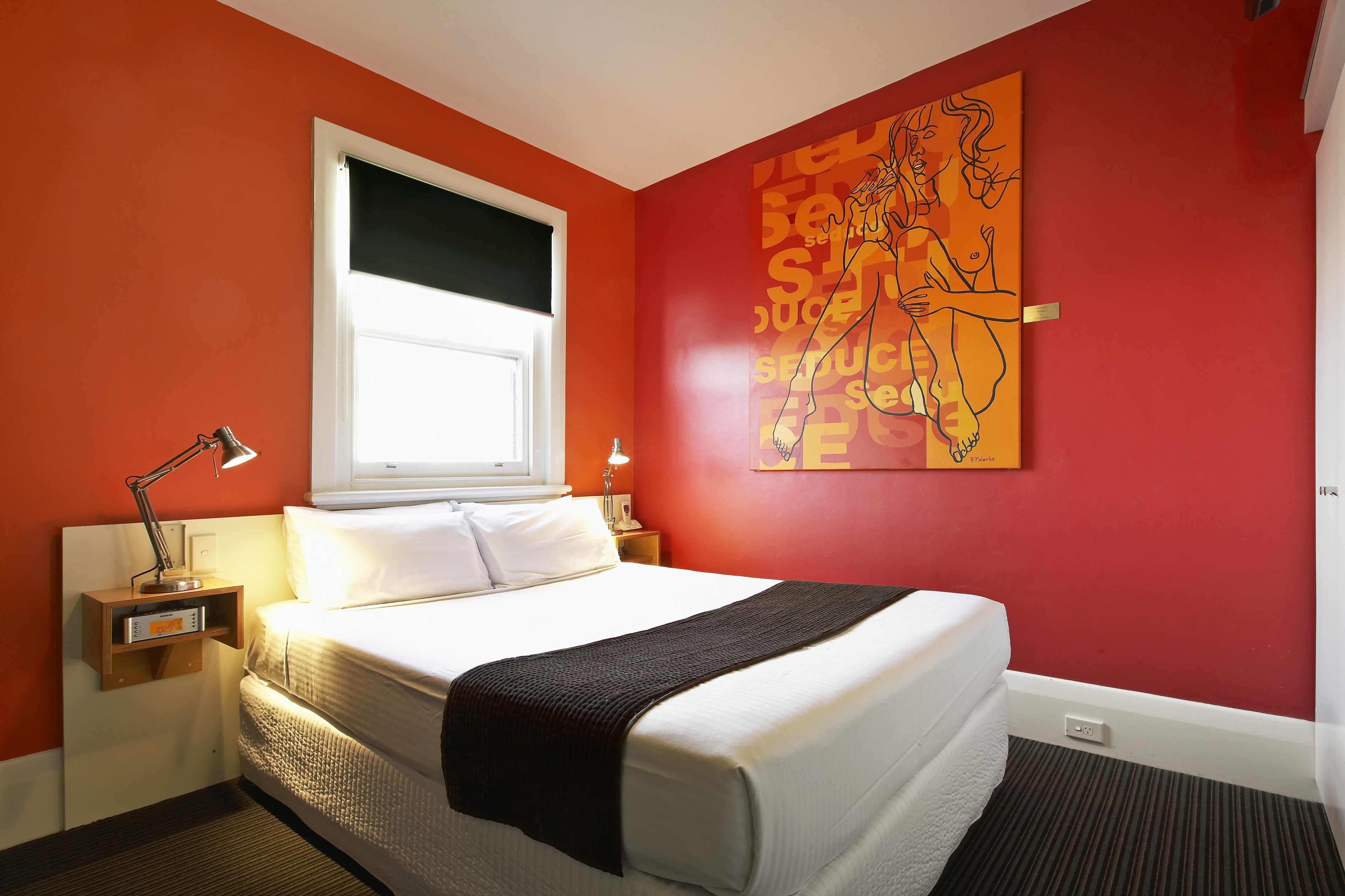 A hotel room with a large bed, a window with blinds, a nightstand with a lamp, and a wall with a colorful abstract painting.