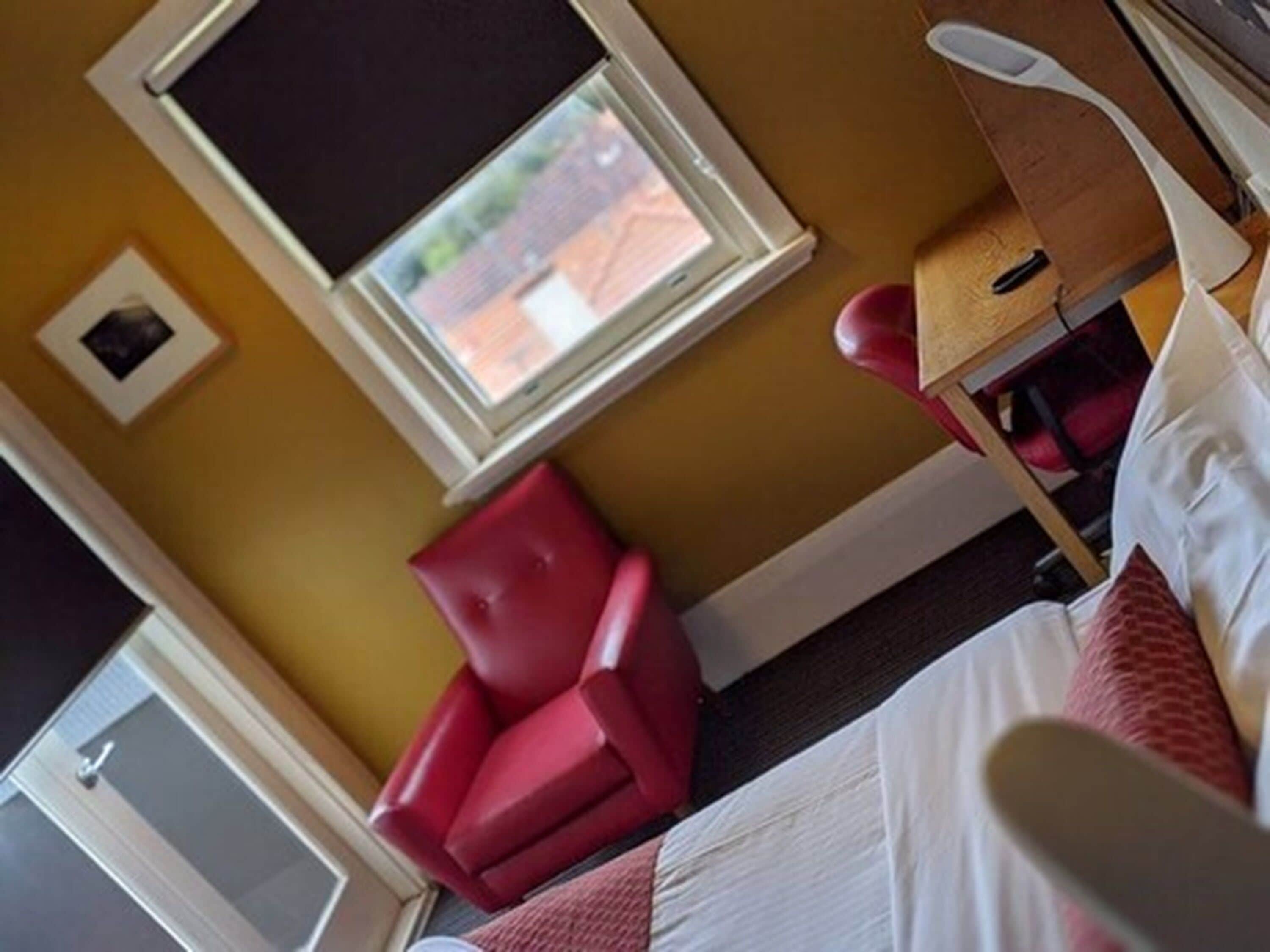 A hotel room with a bed, a red chair, a small table, and a window with a view of the outside.