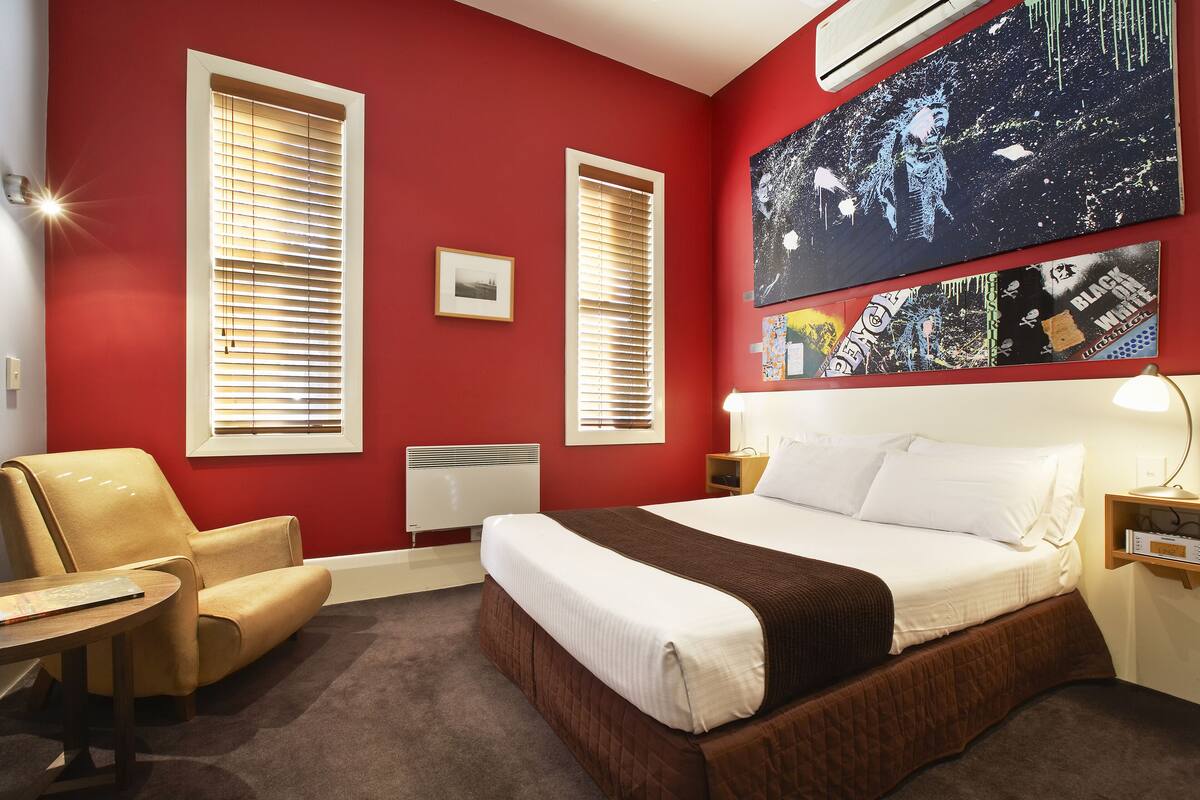 A hotel room with a large bed, a chair, a small table, and two windows with blinds.