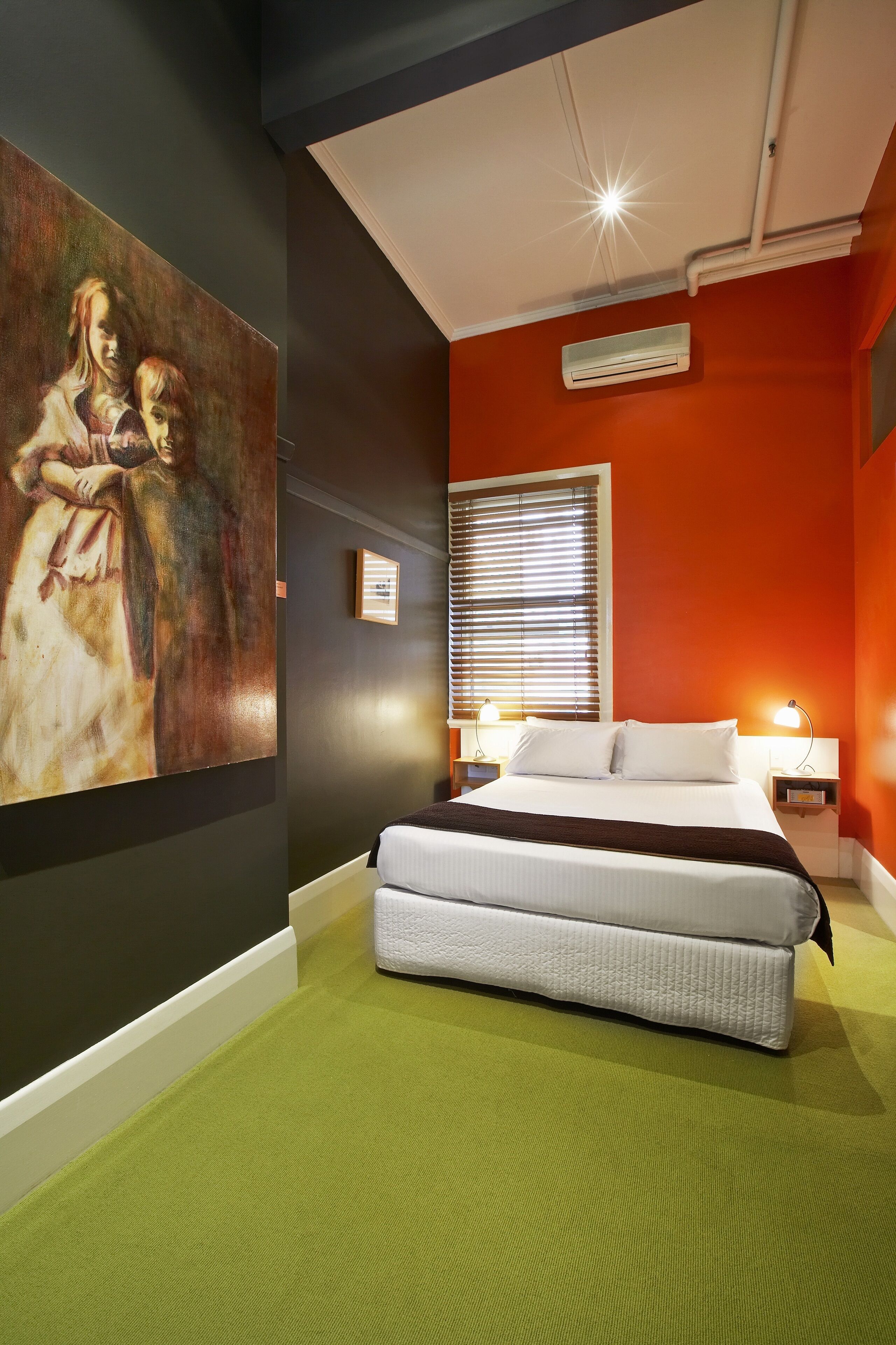 A bedroom with a bed, a painting on the wall, a window with blinds, and an air conditioning unit.
