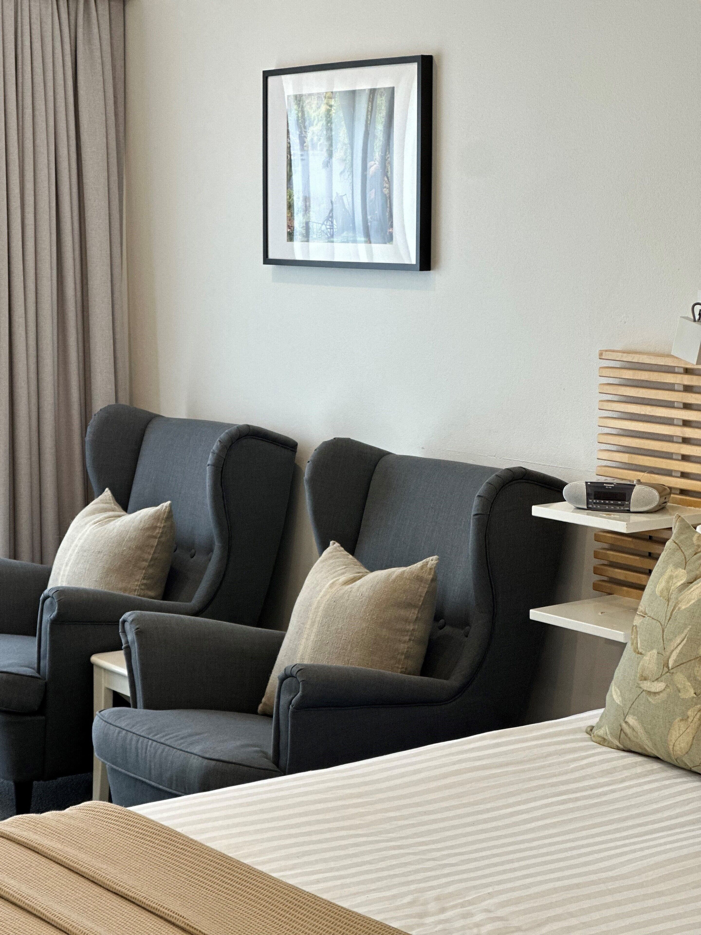 A hotel room with two dark grey armchairs, a bed with a striped bedspread, a framed picture on the wall, and a small table with a radio.