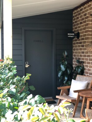 A dark grey door labeled 'ROSIER 1' with a brick wall and a wooden bench with a cushion. - Berrima Bakehouse Motel (Berrima)