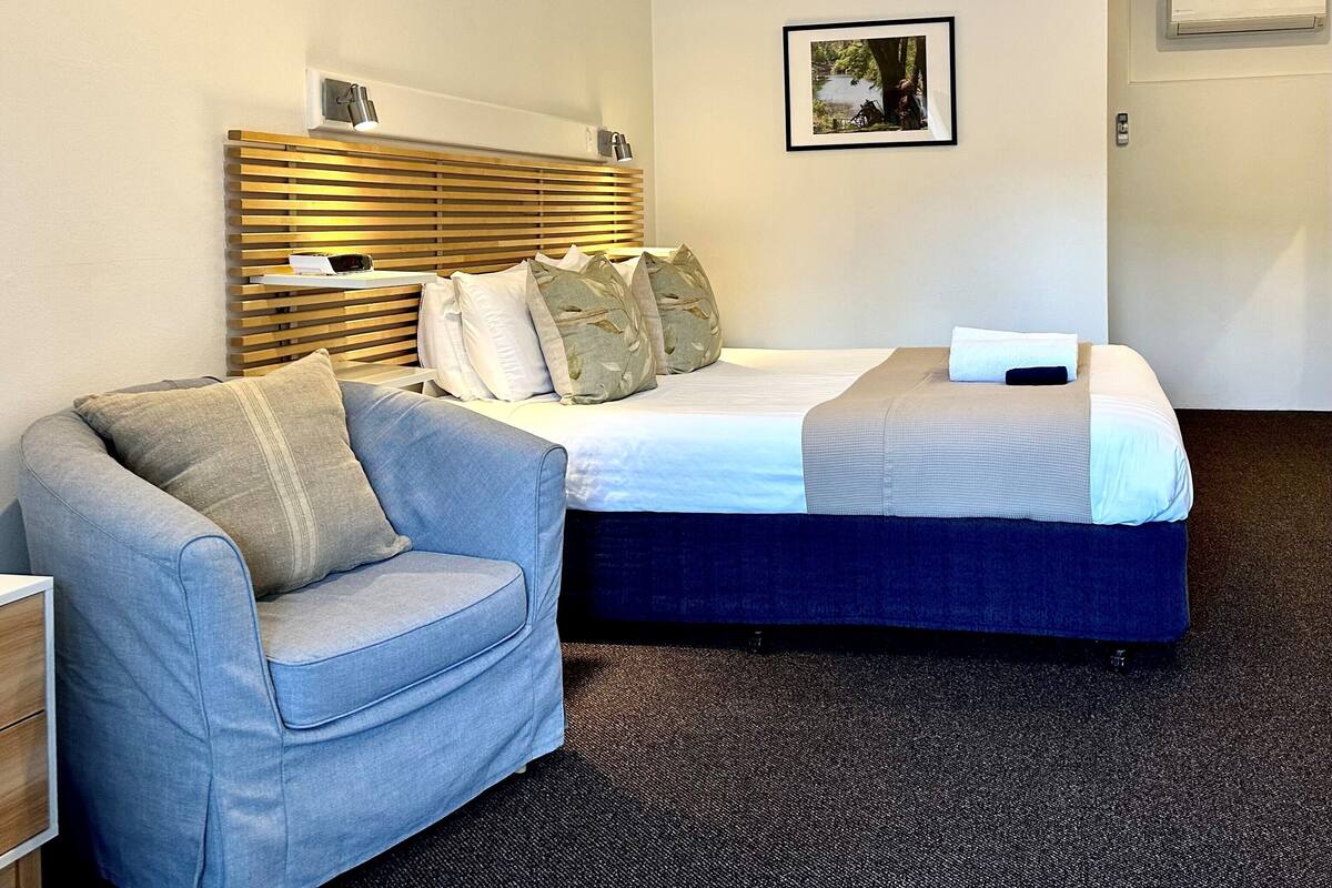 A hotel room with a bed, a chair, a picture on the wall, and an air conditioning unit.